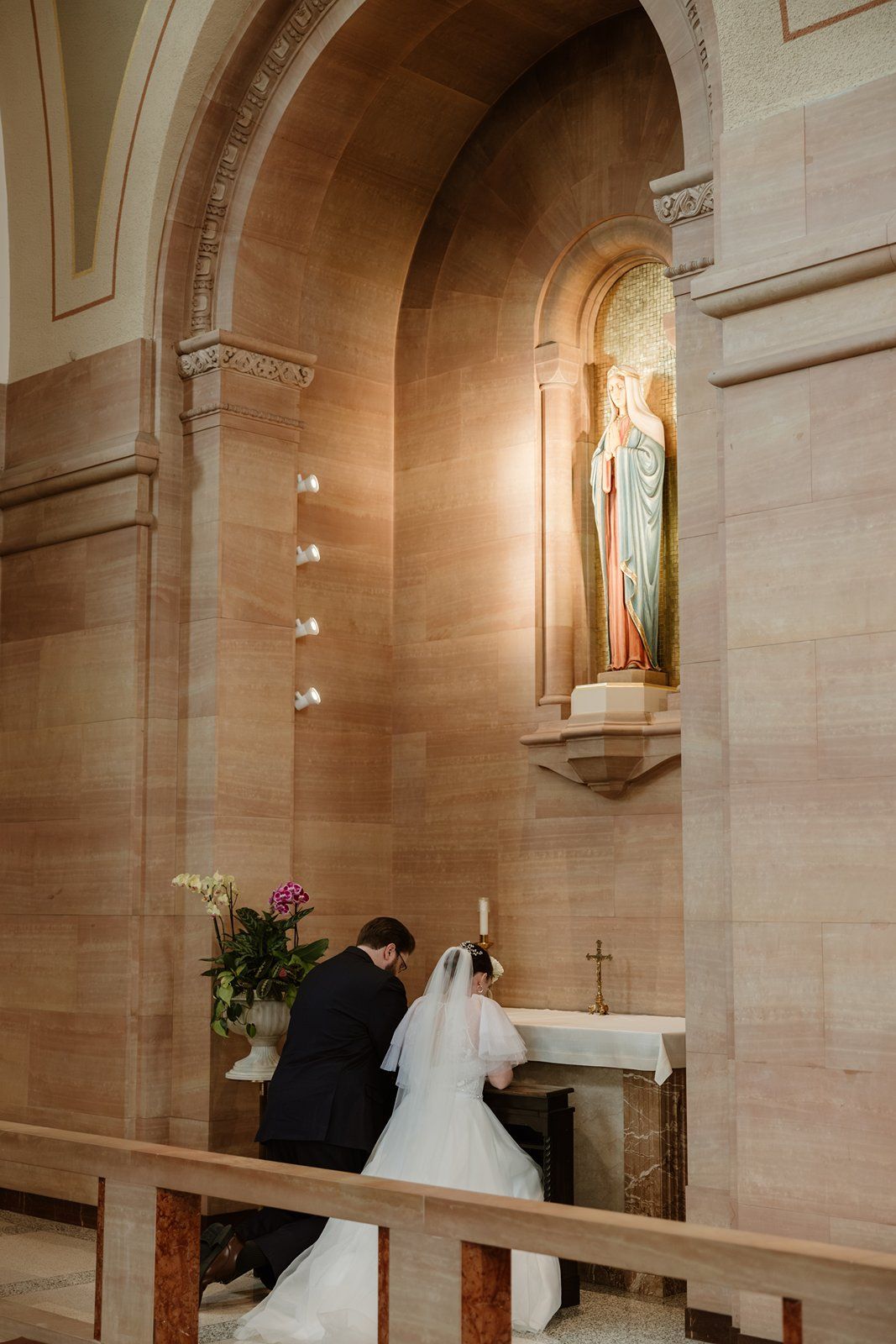 A couple kneels at the altar during a Catholic church wedding ceremony with stained glass windows and stone walls.