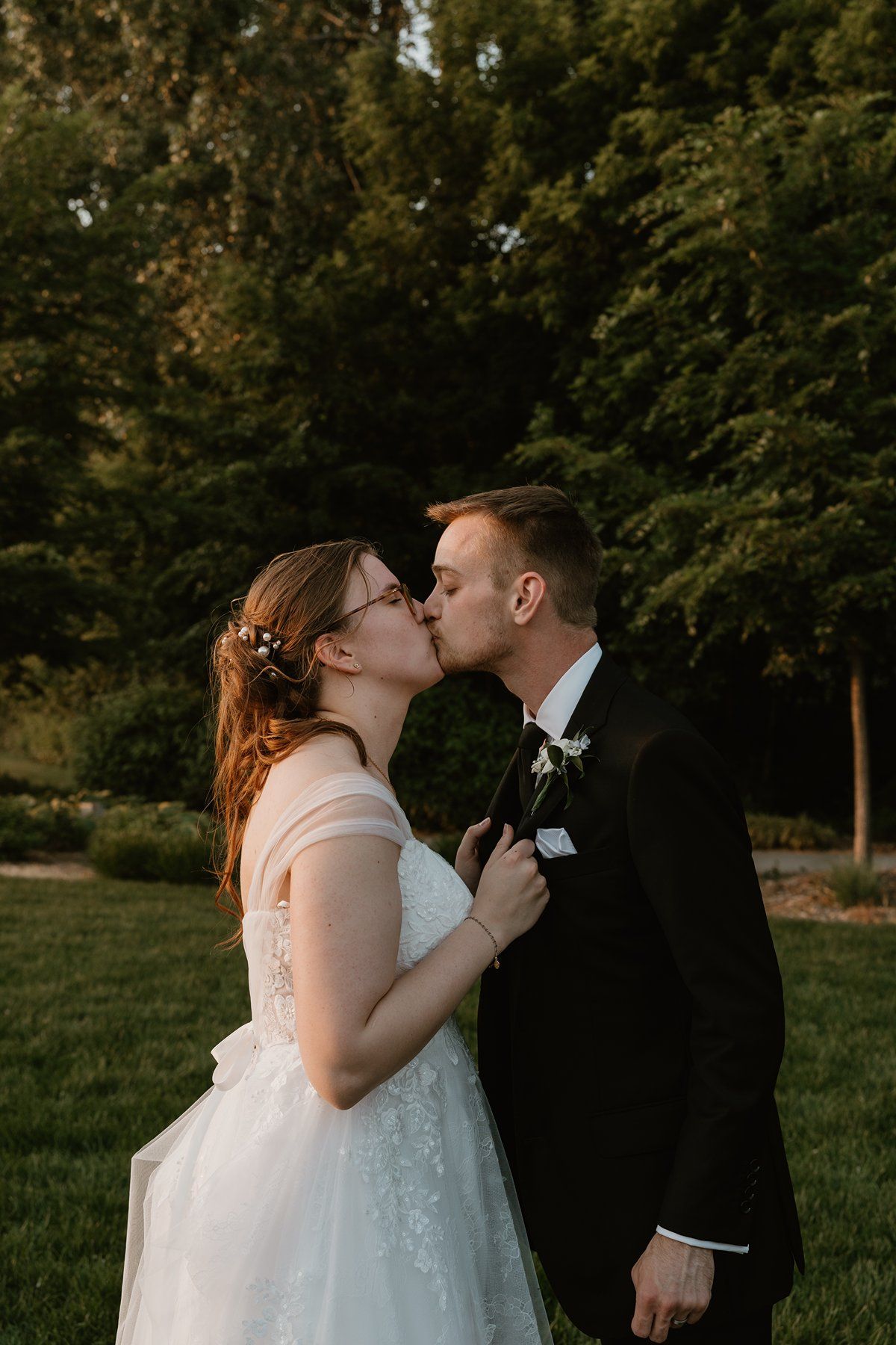 A romantic couple in wedding attire share a passionate kiss while standing in an outdoor garden setting at sunset.