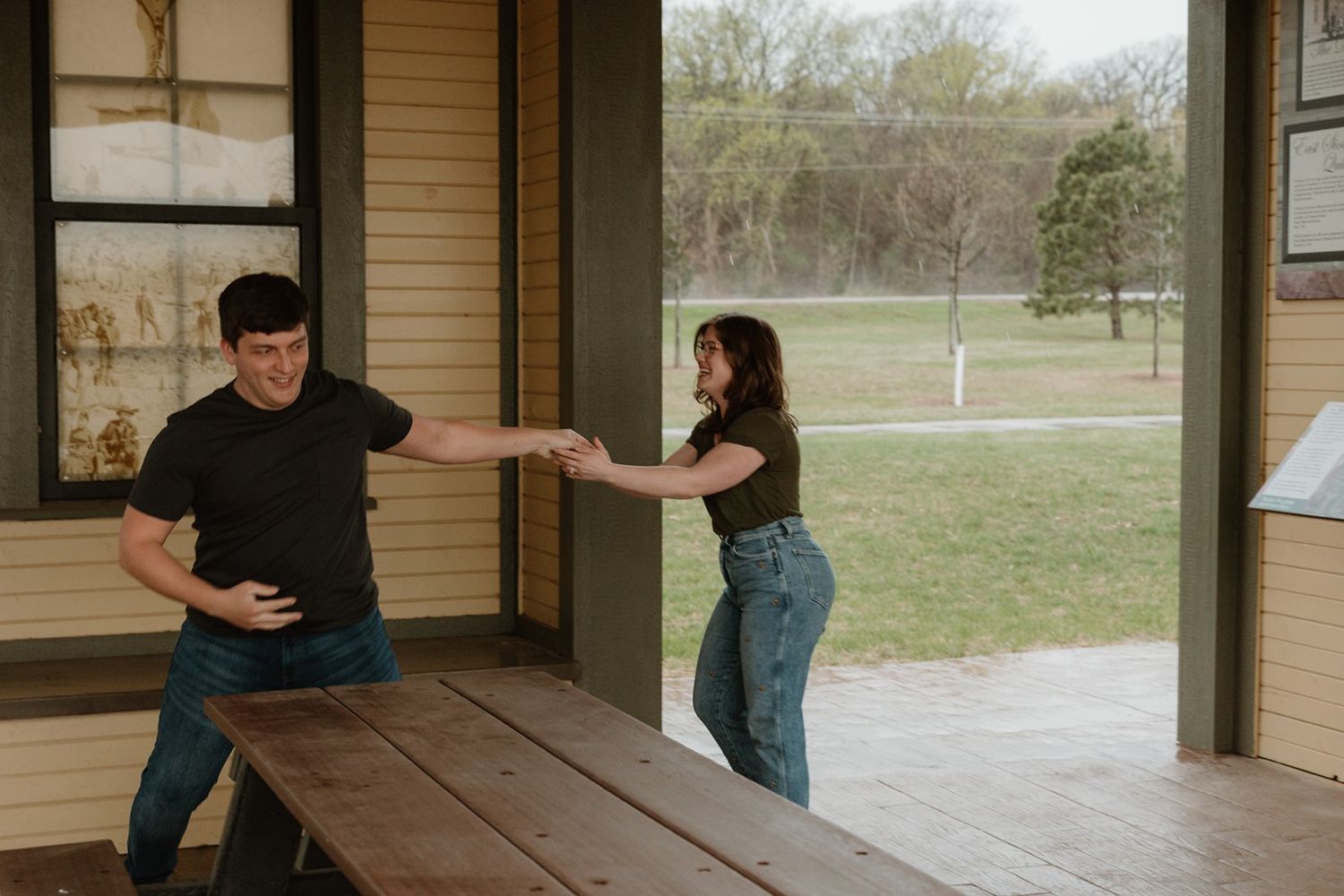 Two people playfully interacting on a covered wooden porch with a grassy field visible in the background.