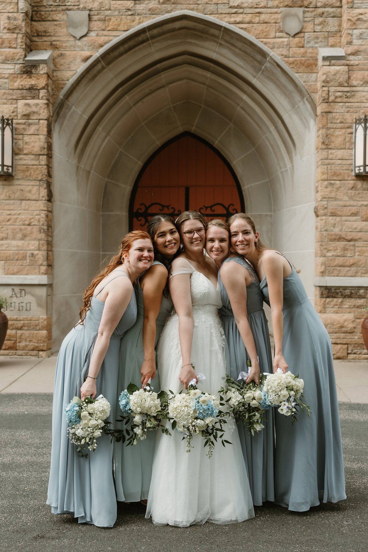 Group in pale blue bridesmaid dresses and white wedding gown pose with white floral bouquets in front of stone church archway.