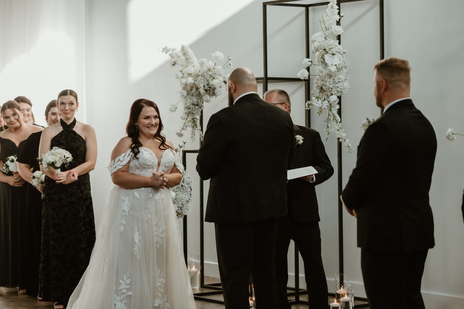 A couple exchanges vows during indoor wedding ceremony with white floral arch and wedding party in dark attire.