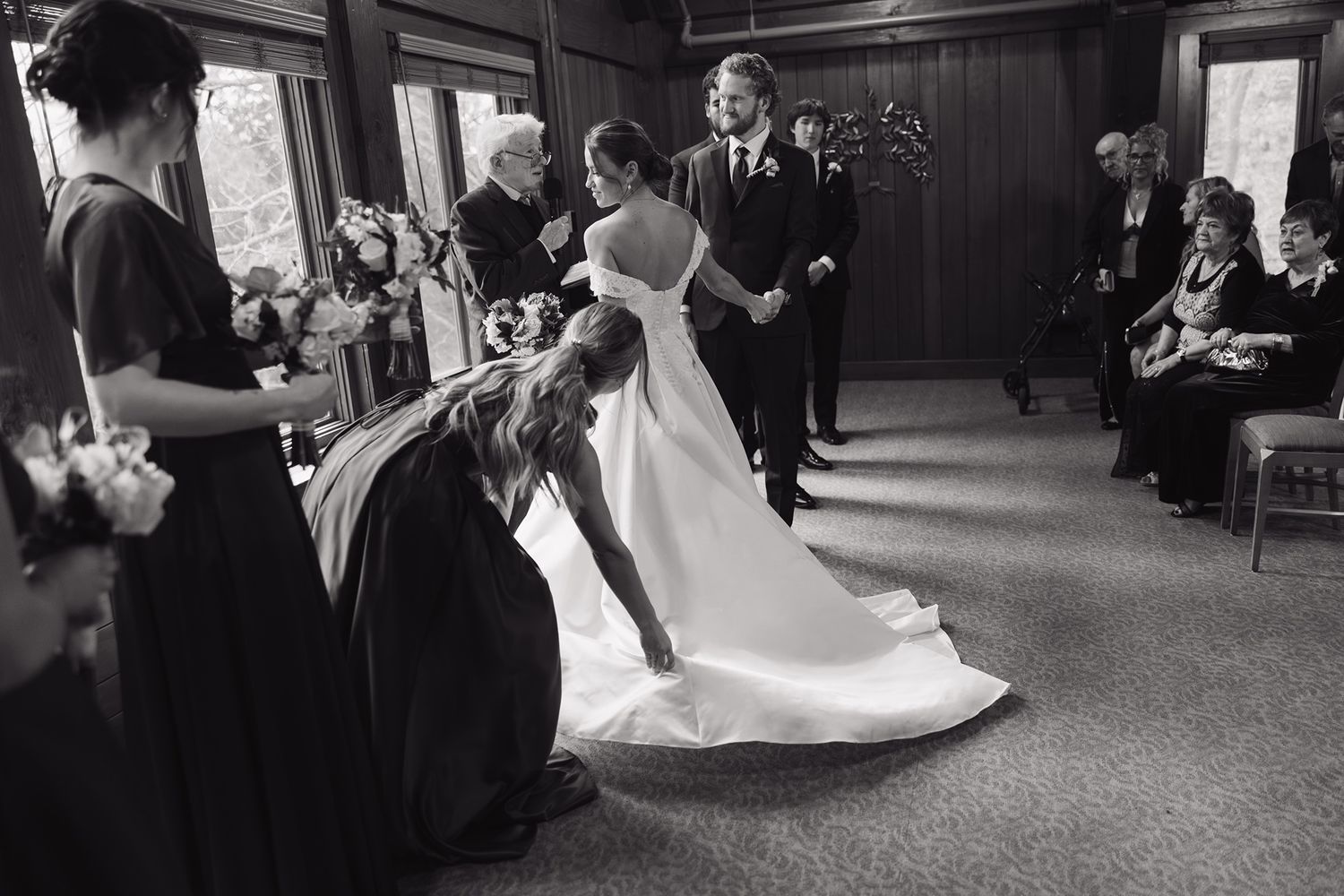 Black and white wedding photo of bride having her dress arranged by bridesmaid during indoor ceremony.