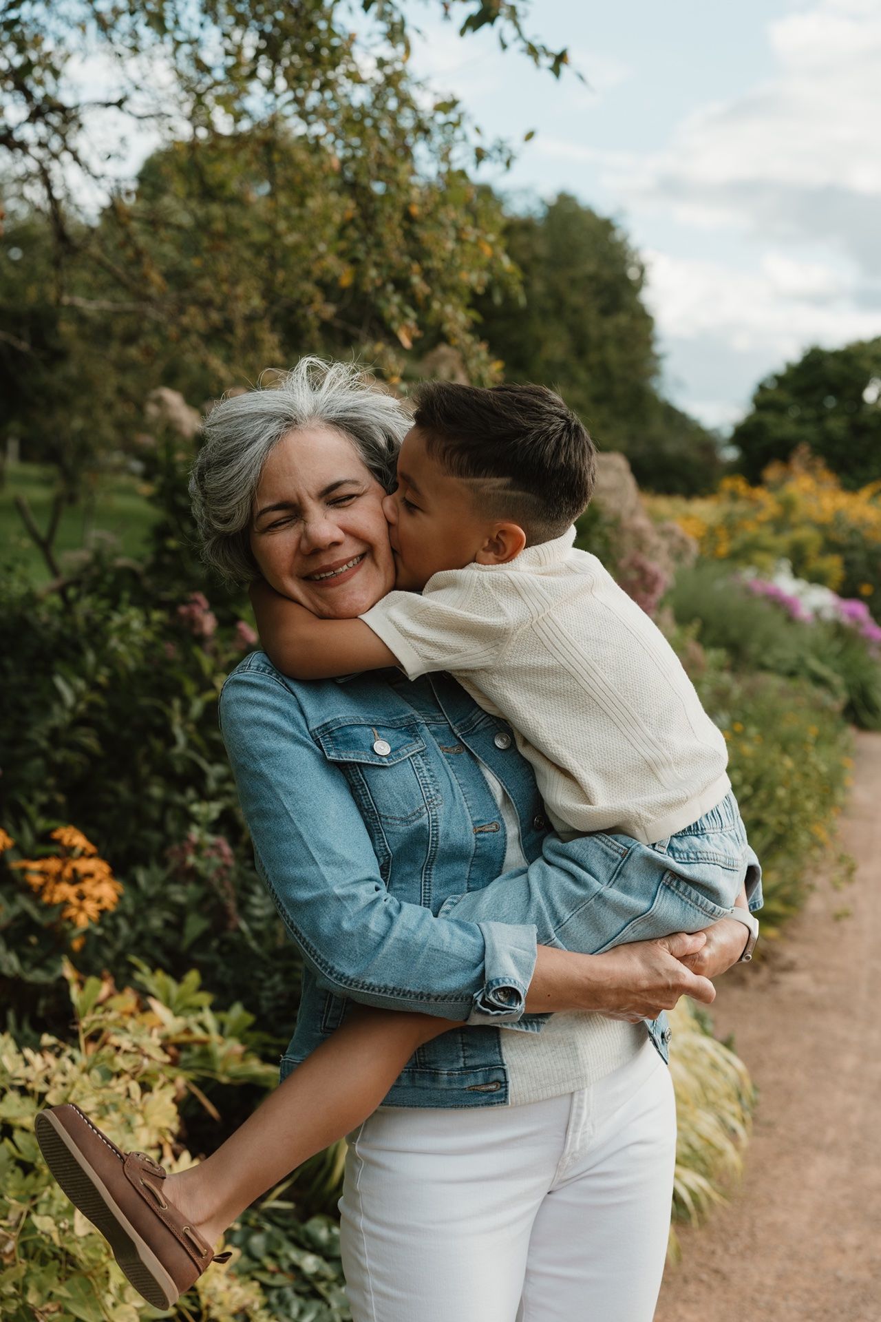Smiling grandmother with gray hair holds young boy in garden, sharing a tender cheek kiss outdoors.