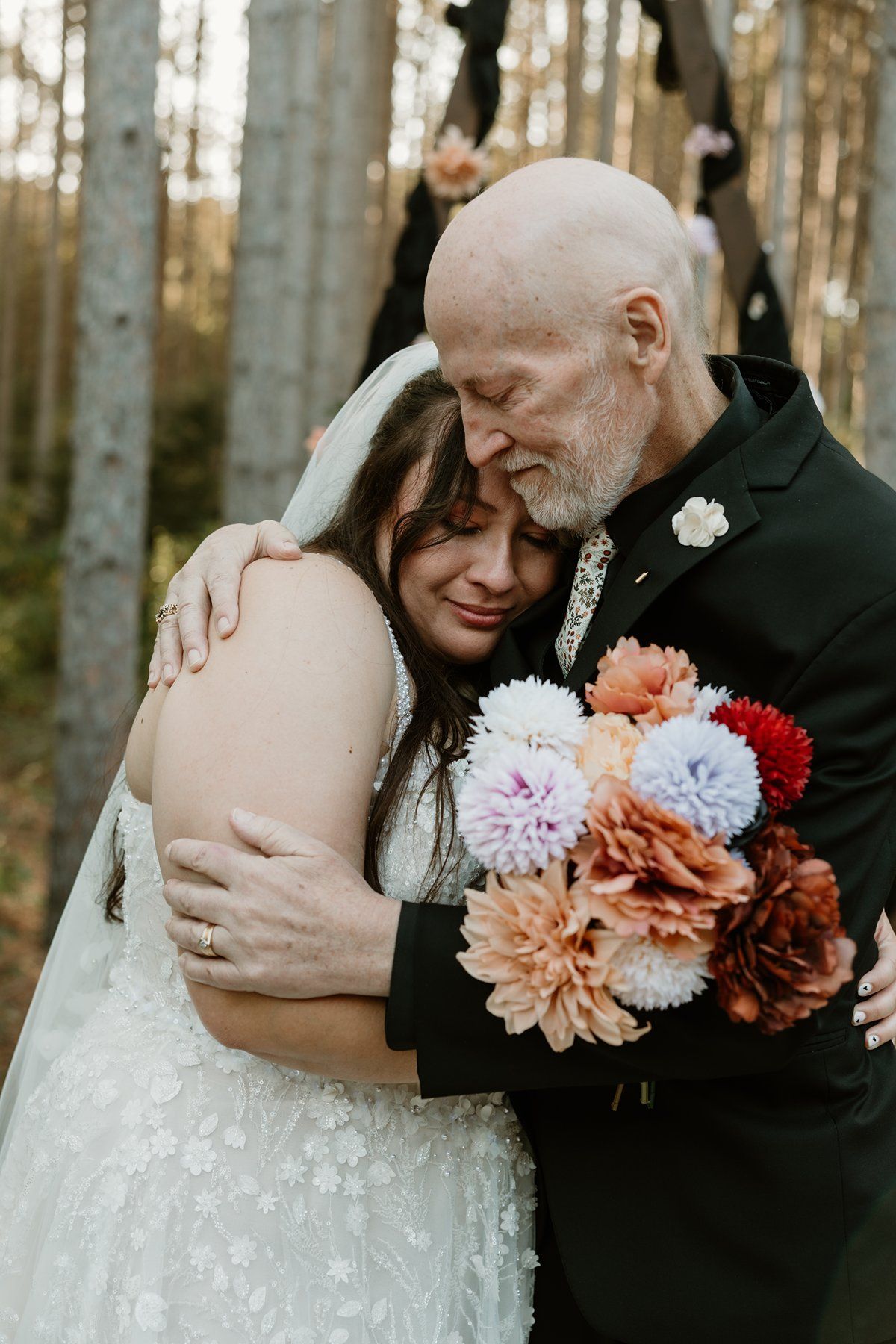 Emotional bride and her father embracing in a forest wedding ceremony, holding colorful dahlia bouquet.