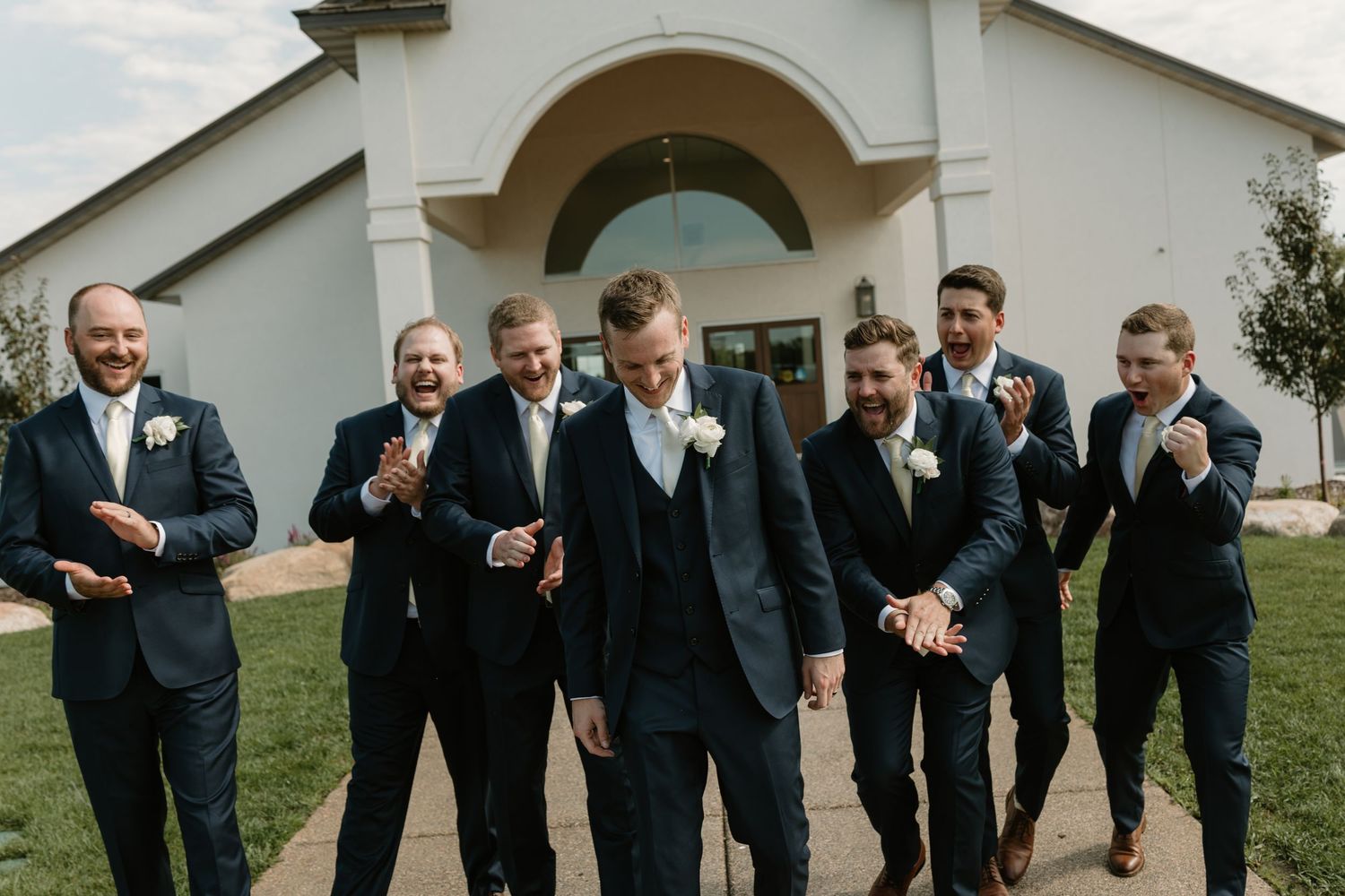 A line of groomsmen in black suits perform a fun choreographed dance move outdoors at a wedding celebration.
