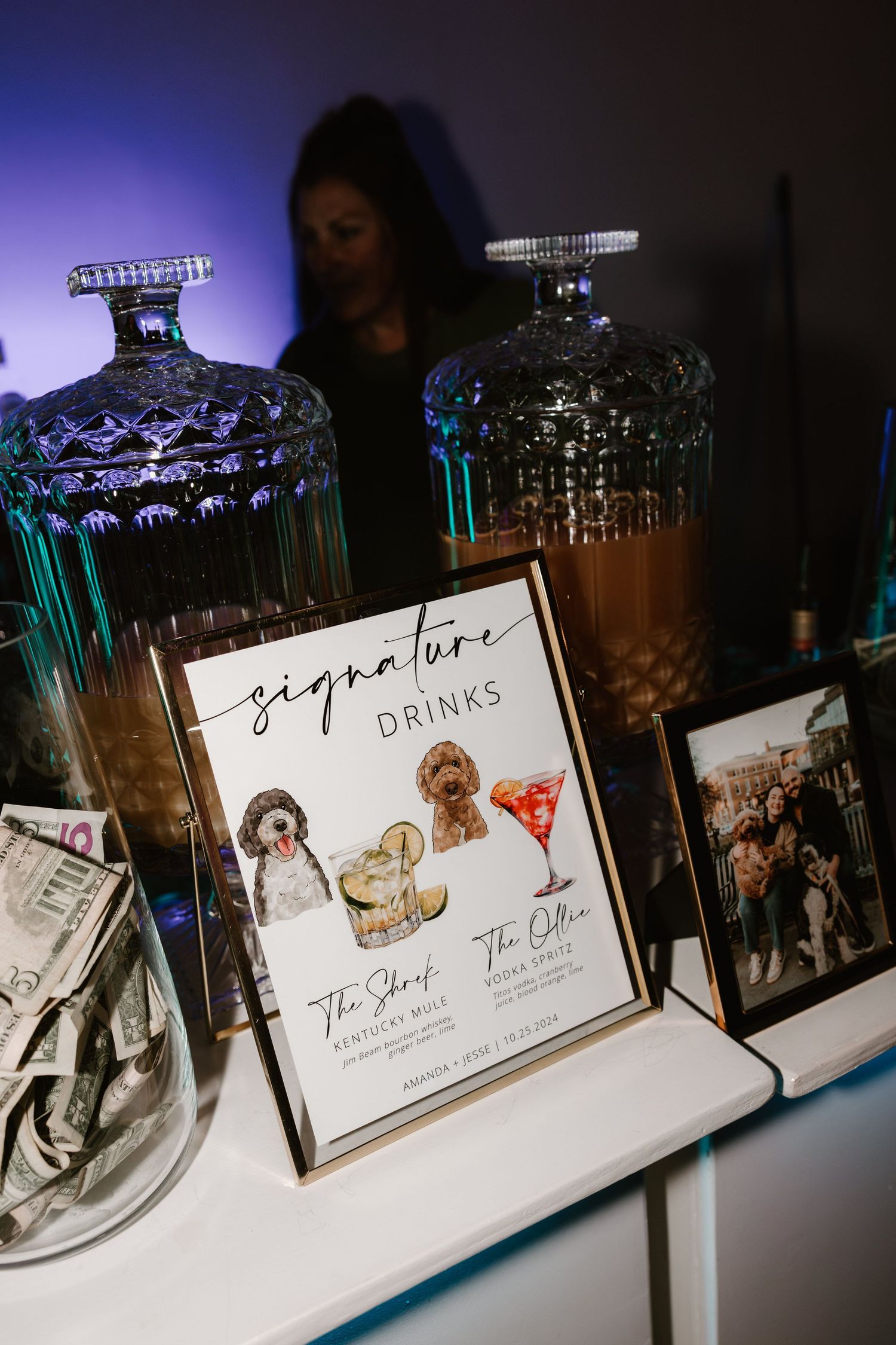 Signature drinks menu displayed in gold frame next to decorative glass decanters with purple and blue lighting at event bar.