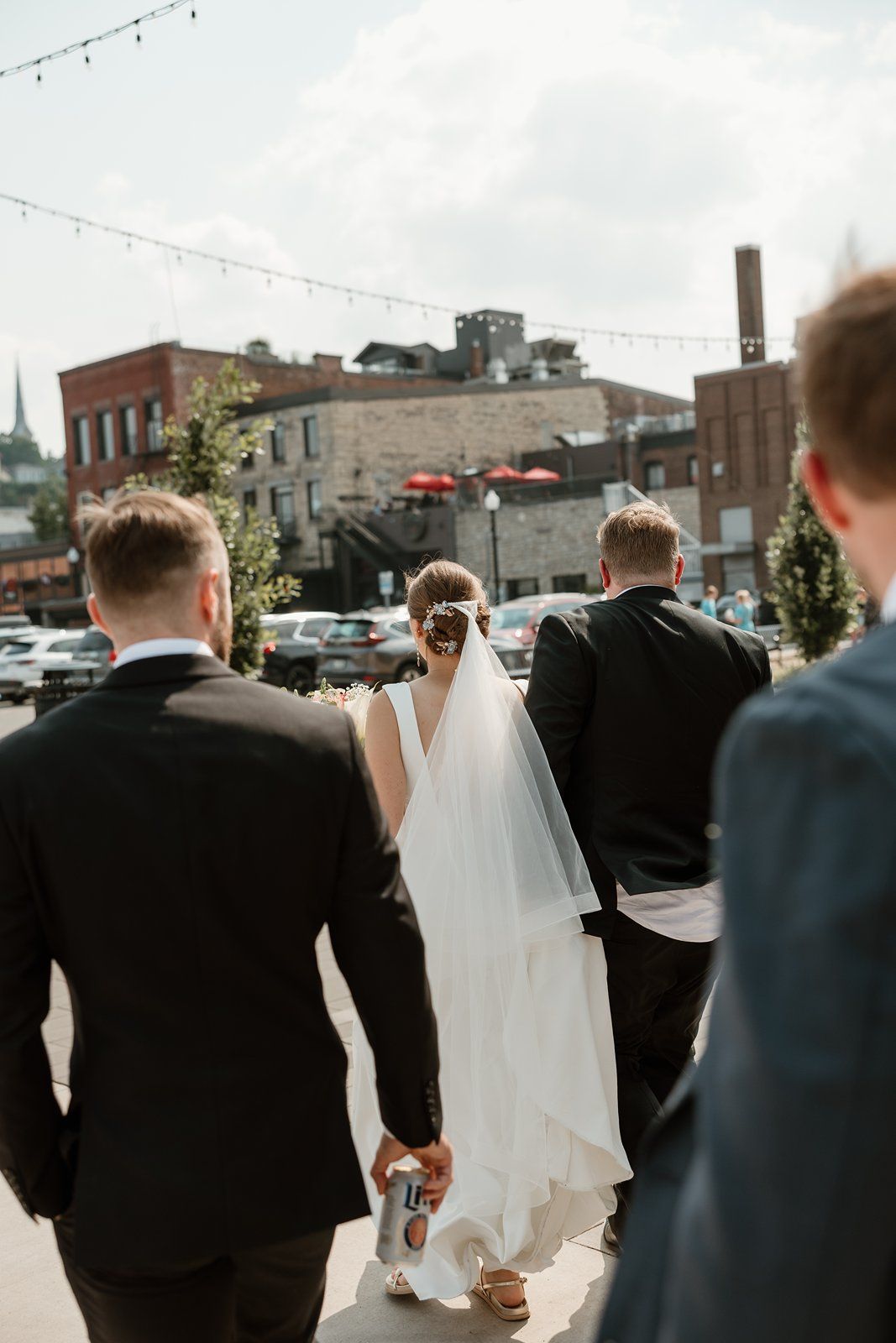A wedding party walks down a city street with string lights overhead and brick buildings in the background.