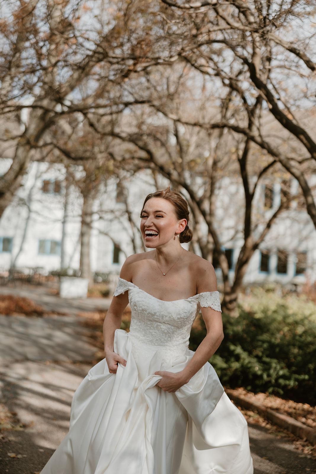 Bride in off-shoulder gown laughing joyfully in autumn garden setting.
