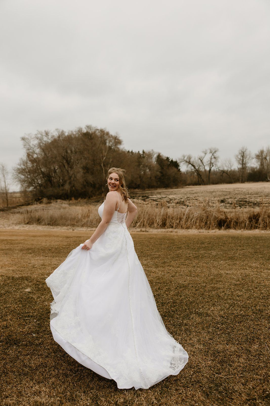 A person in a flowing white wedding dress twirls in a rustic outdoor field on an overcast autumn day.