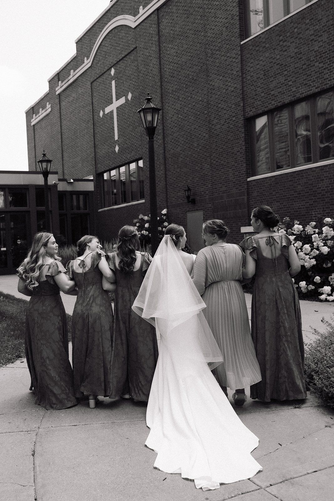 A bride in a flowing white gown and bridesmaids in long dresses gather outside a brick church with a cross on the wall.