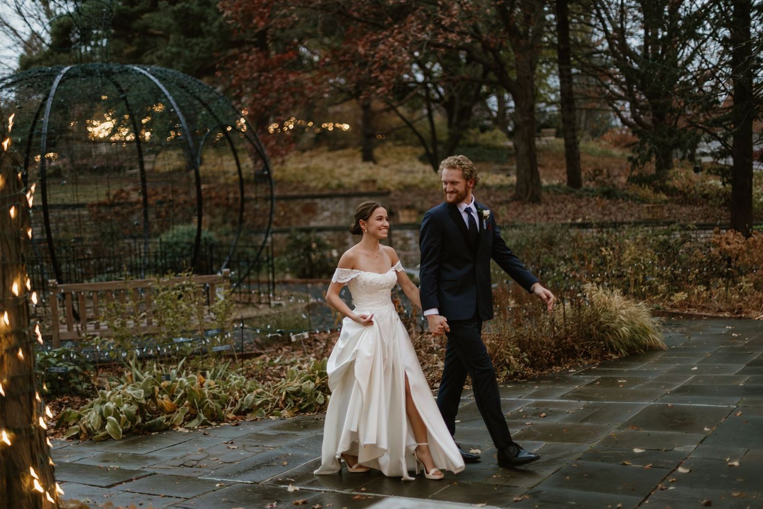 A couple walks under an illuminated garden arch during their autumn evening wedding.