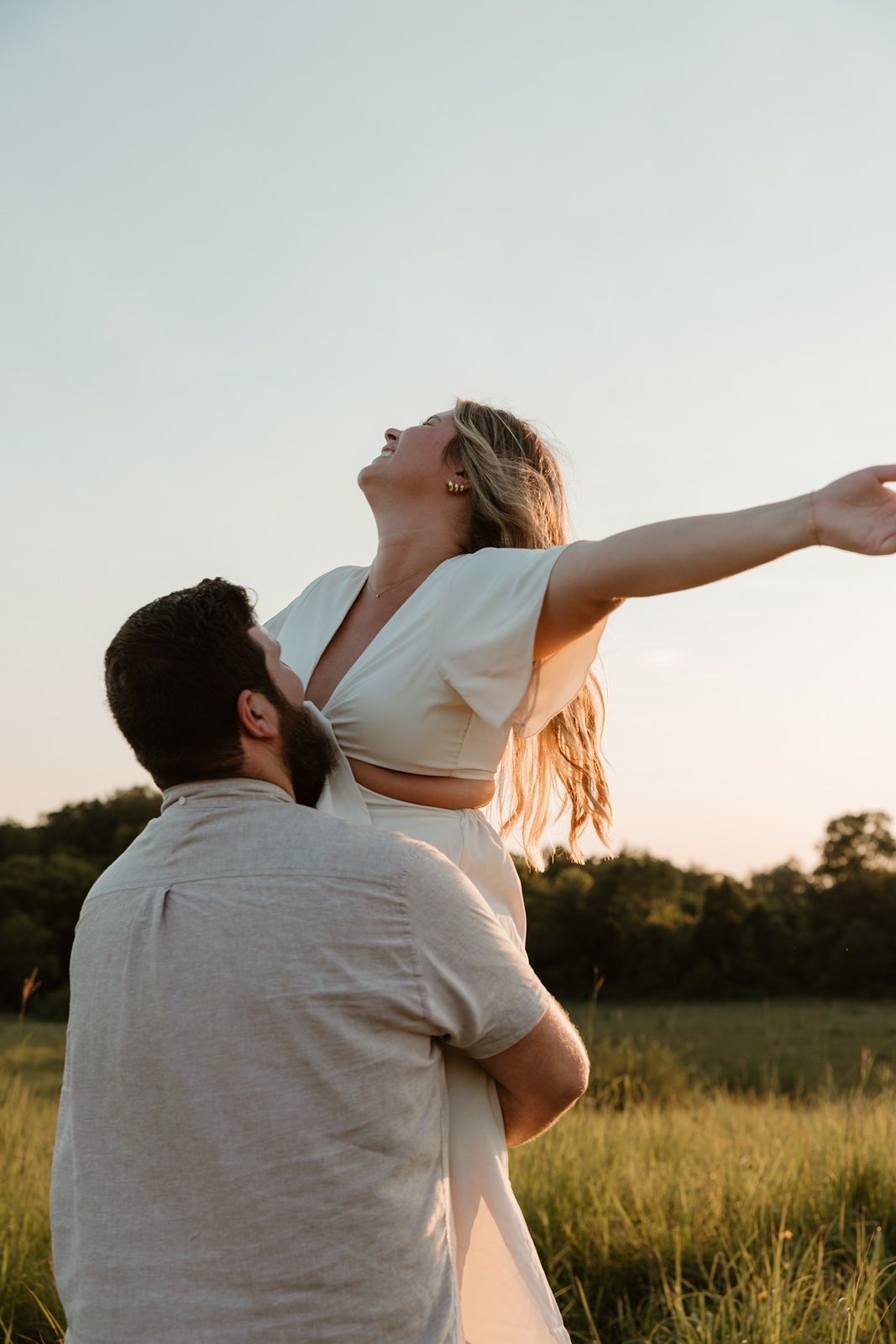 A joyful couple embraces in a sunlit field at golden hour, woman spreading arms wide with head tilted back in bliss.