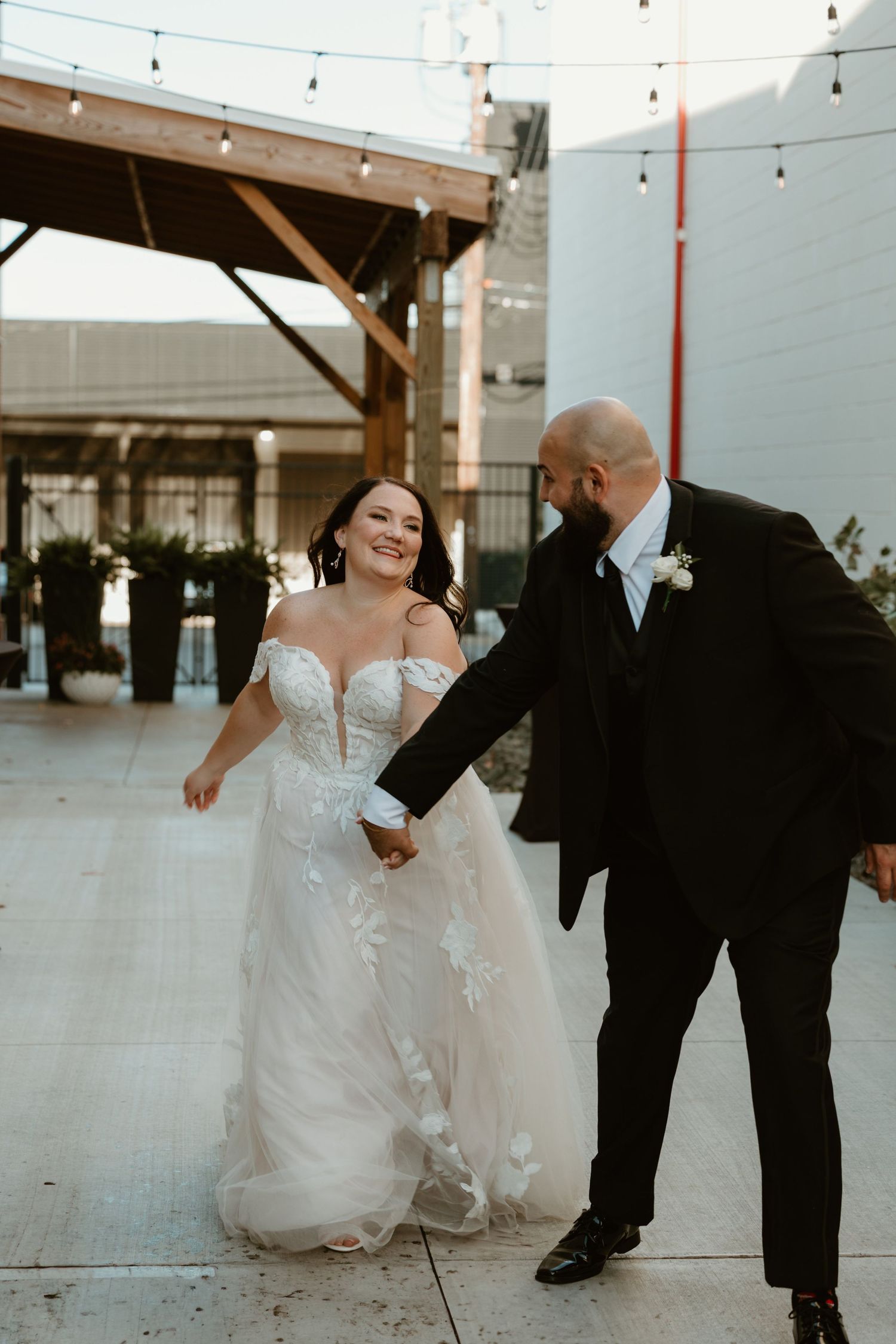 A newlywed couple dances and laughs together on an outdoor patio decorated with string lights.