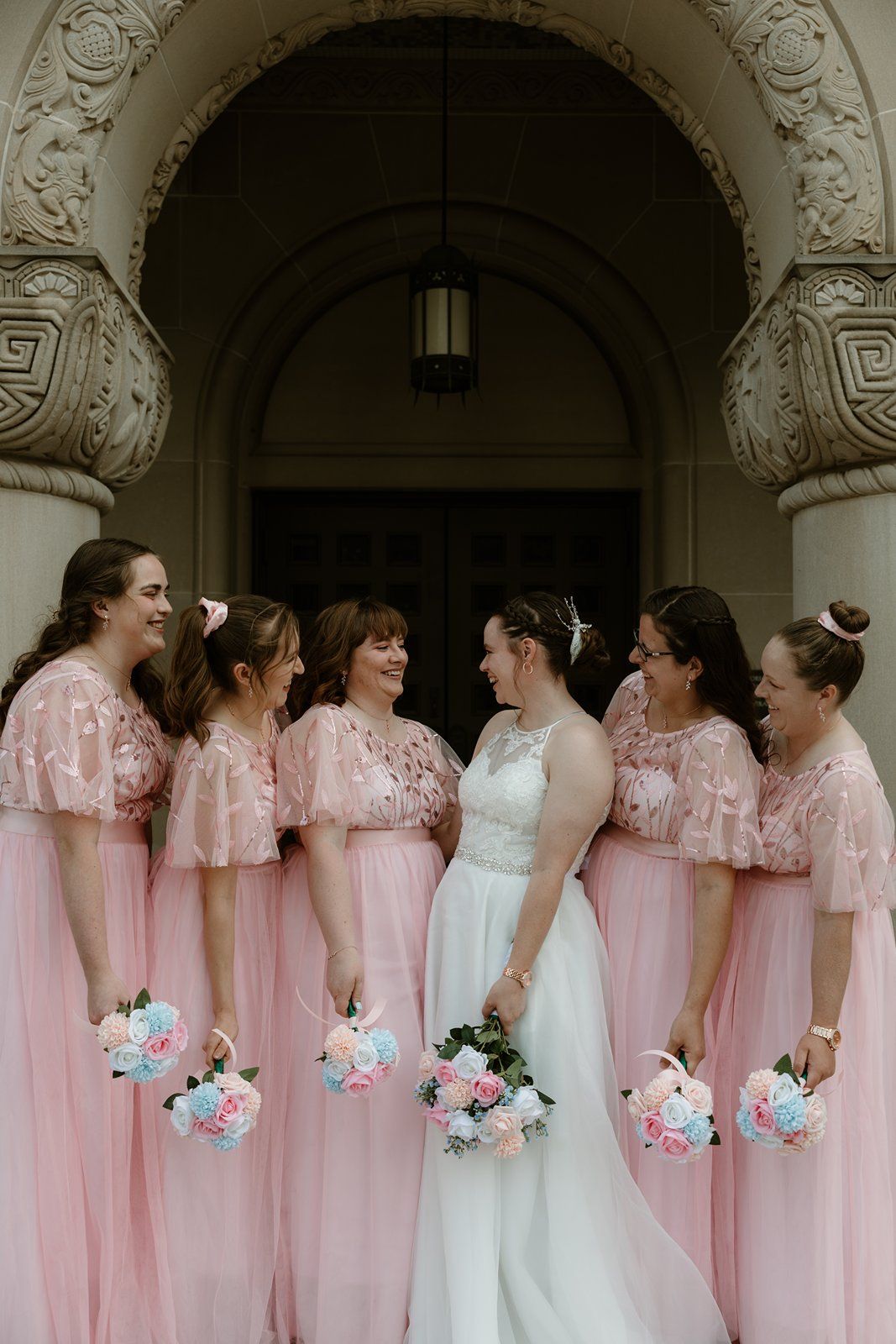 Bridesmaids in flowing pink dresses with lace details gather around bride in white gown holding pastel bouquets.