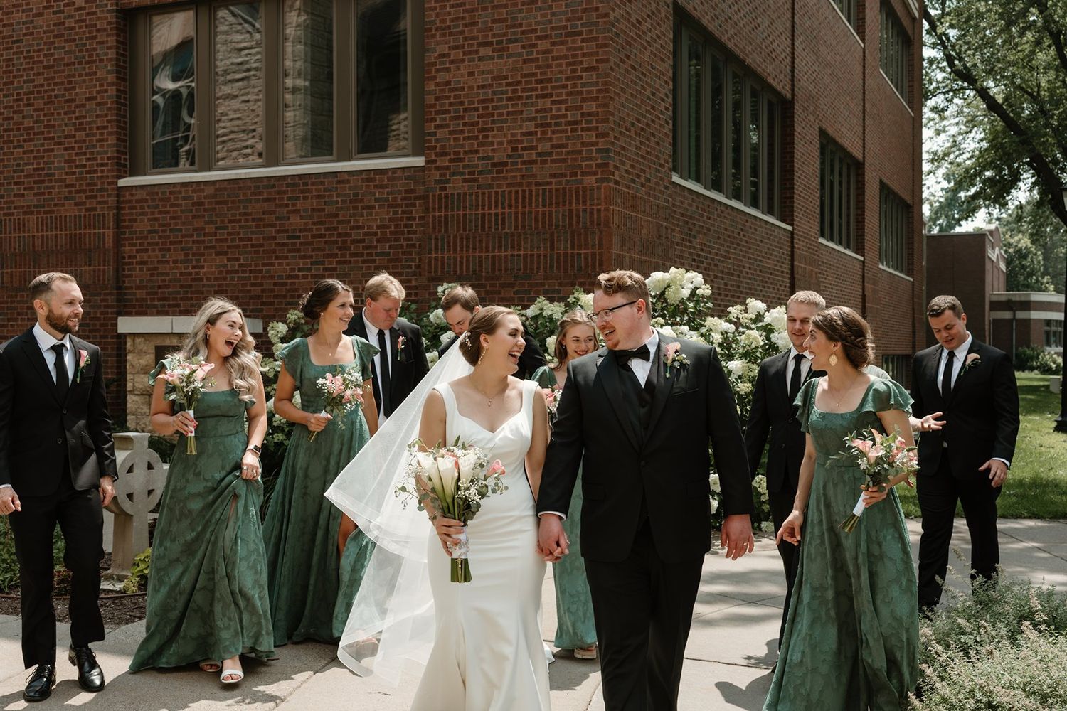Wedding group poses in front of brick church building with bridesmaids in sage green dresses.