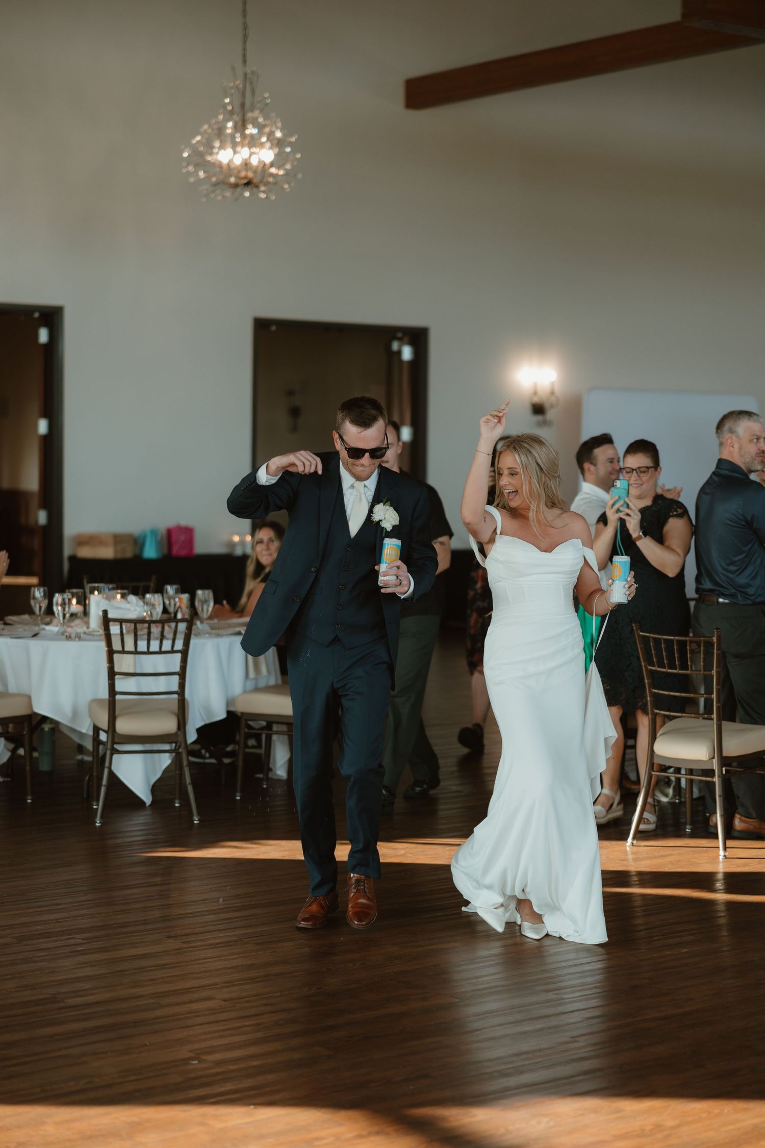 Wedding guests dance together on a dark wooden floor under crystal chandeliers during an evening reception celebration.