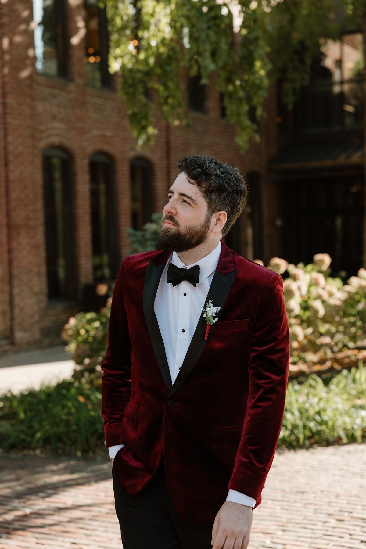 Groom in burgundy velvet tuxedo jacket with bow tie and boutonniere poses outdoors at brick building venue.