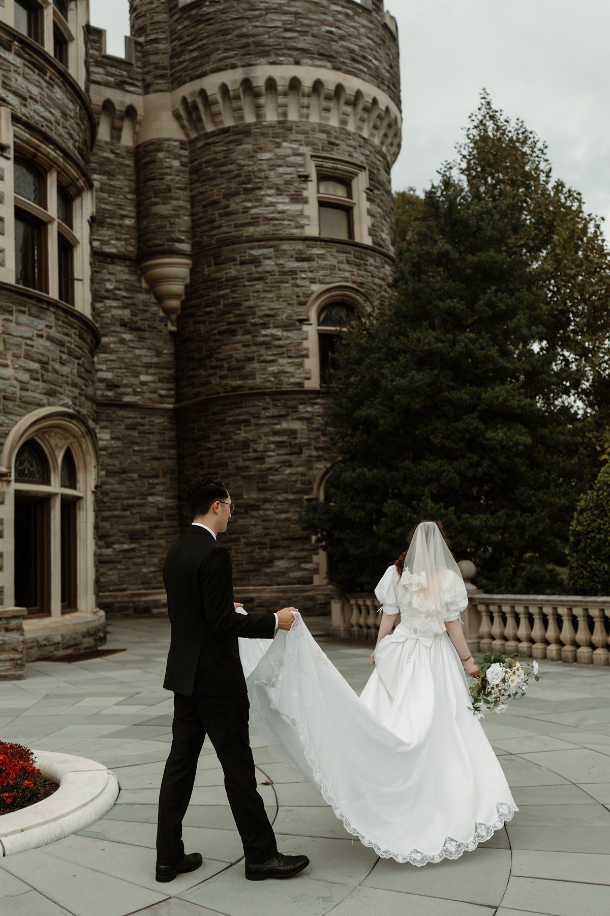 Bride and groom walk toward a stone castle, groom holding wedding dress train, bride carrying bouquet.