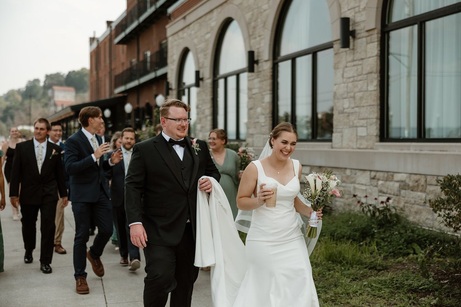 Bride and groom walk together with their wedding party along a rustic stone building exterior.