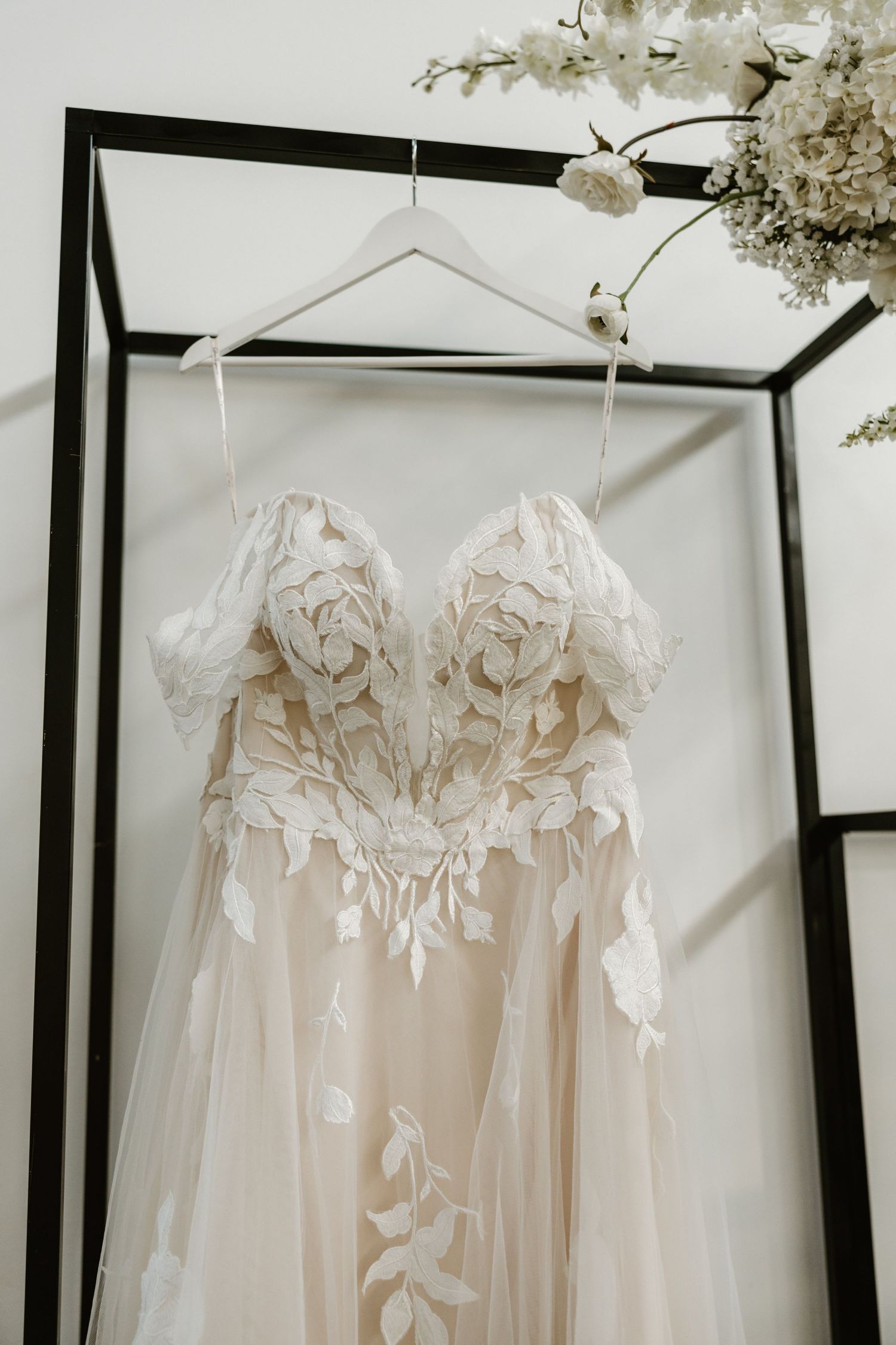 Elegant white wedding dress with delicate lace bodice and sheer tulle skirt hangs on display against neutral backdrop.