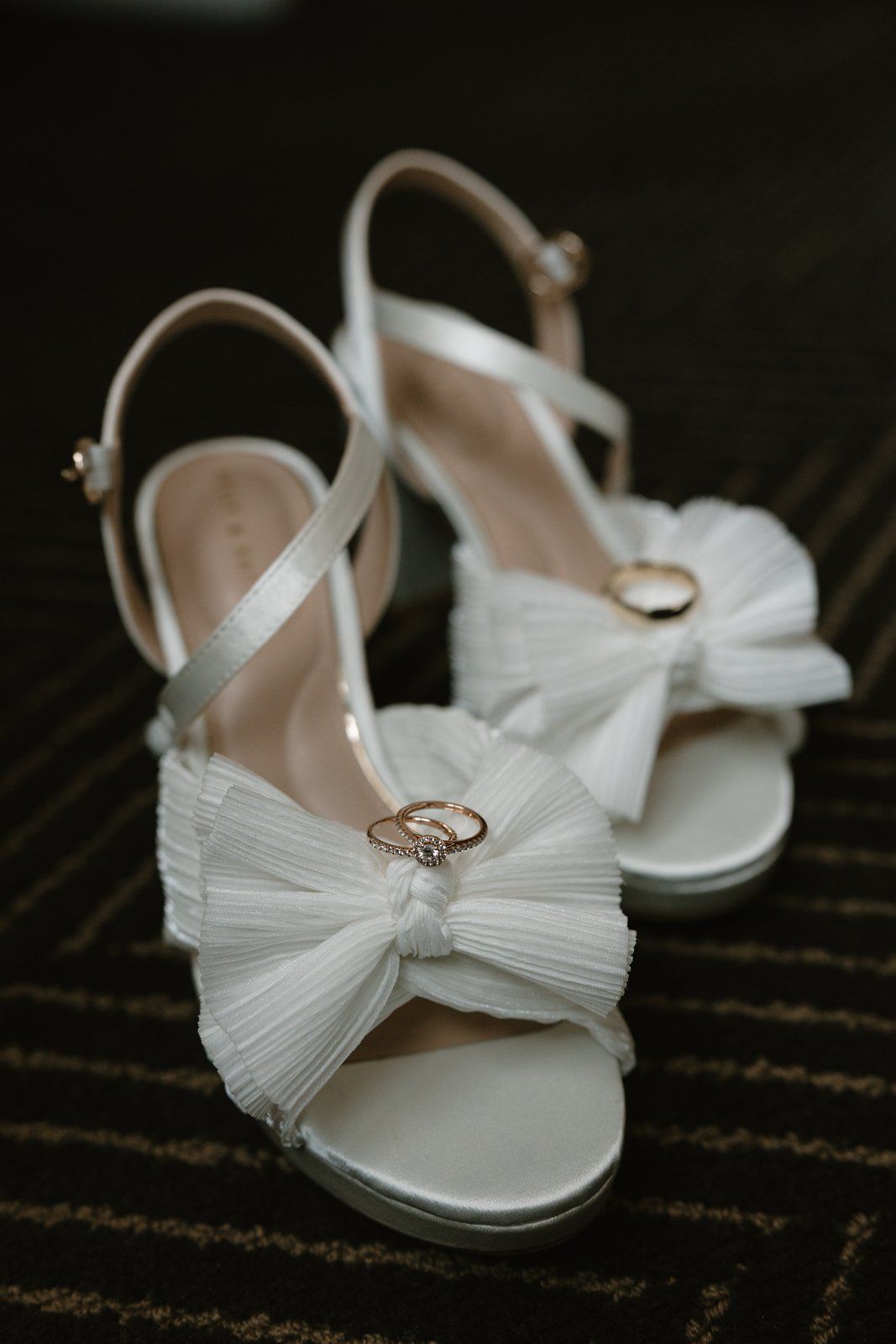 White bridal shoes with tulle bows and a delicate engagement ring displayed on a dark textured surface.