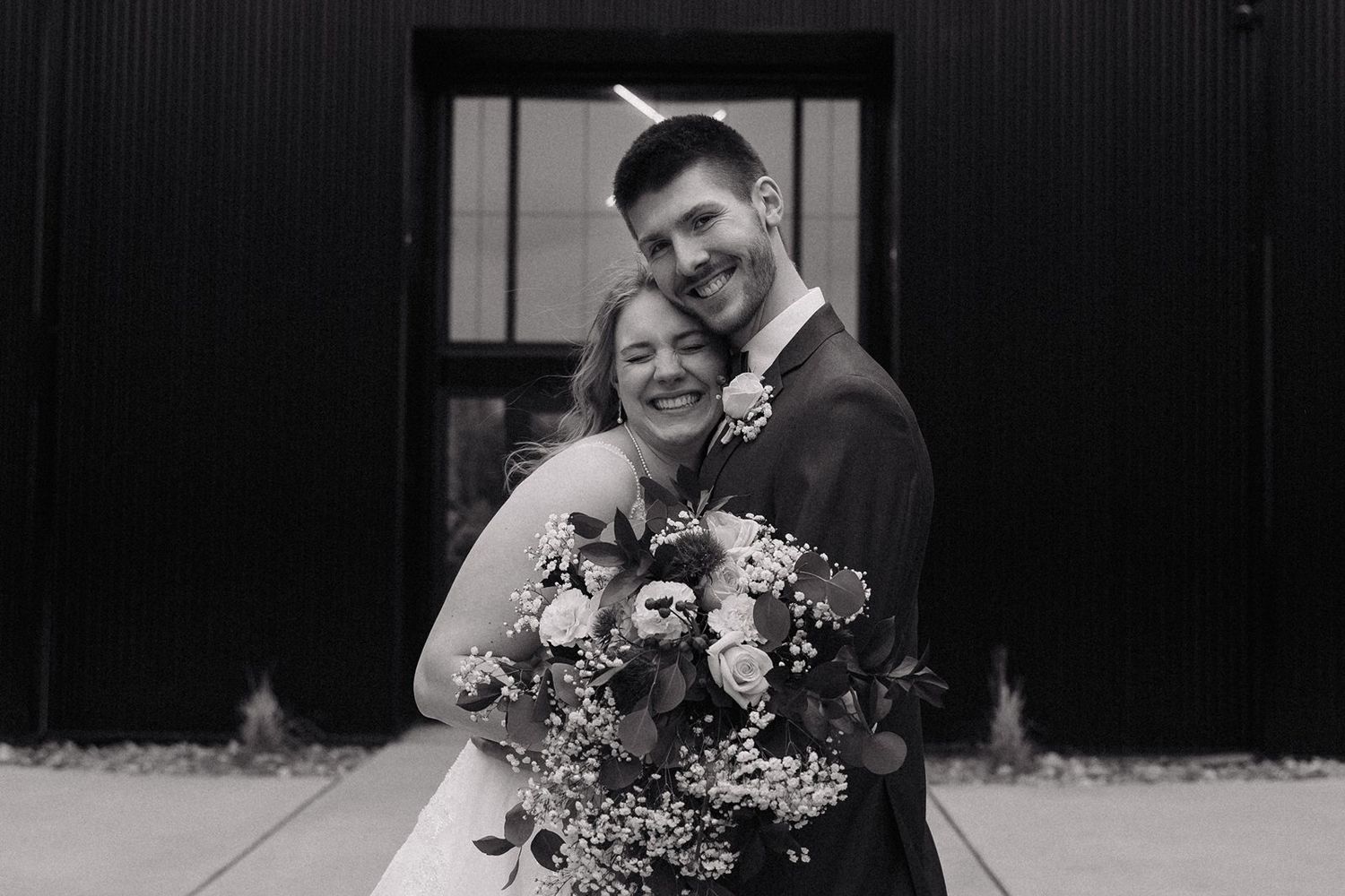 A black and white wedding photo showing a joyful couple embracing while holding a vibrant floral bouquet.