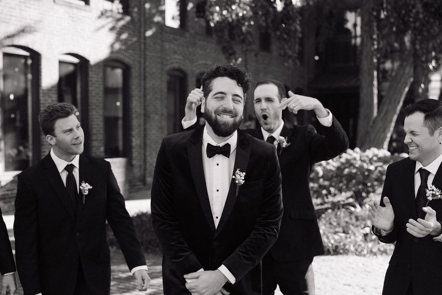 Groom in tuxedo smiles as groomsmen celebrate behind him in black and white wedding photo.