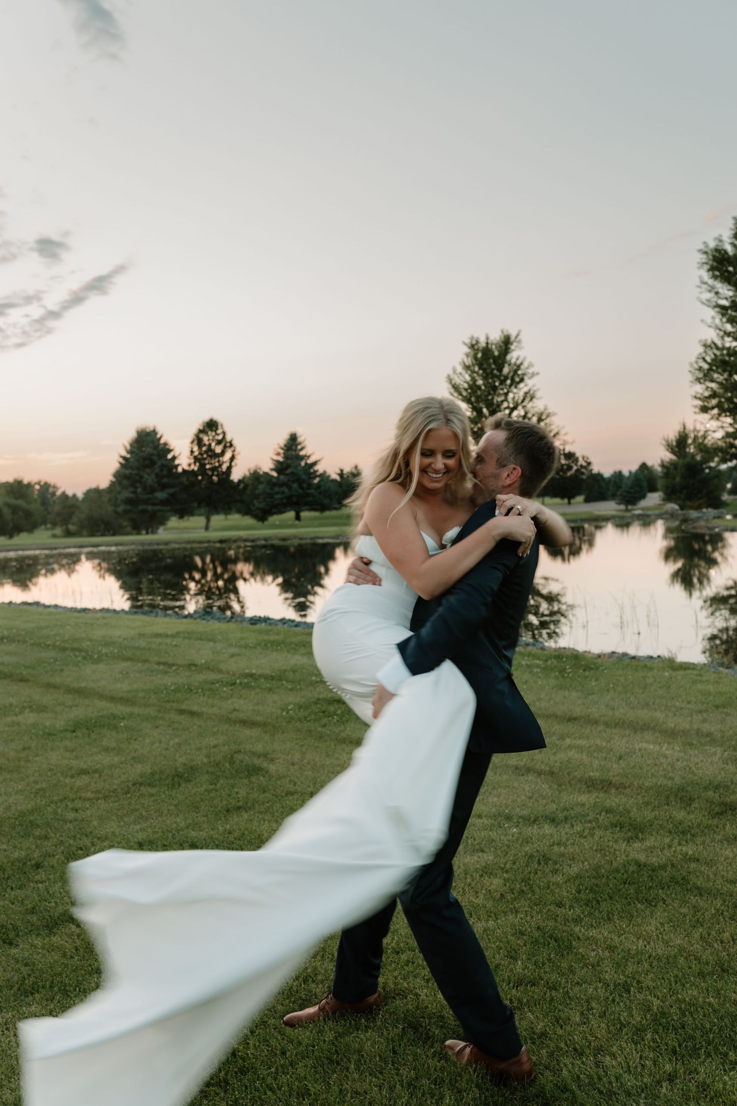 A couple in wedding attire playfully dance and embrace on a grassy lawn near a pond at sunset.