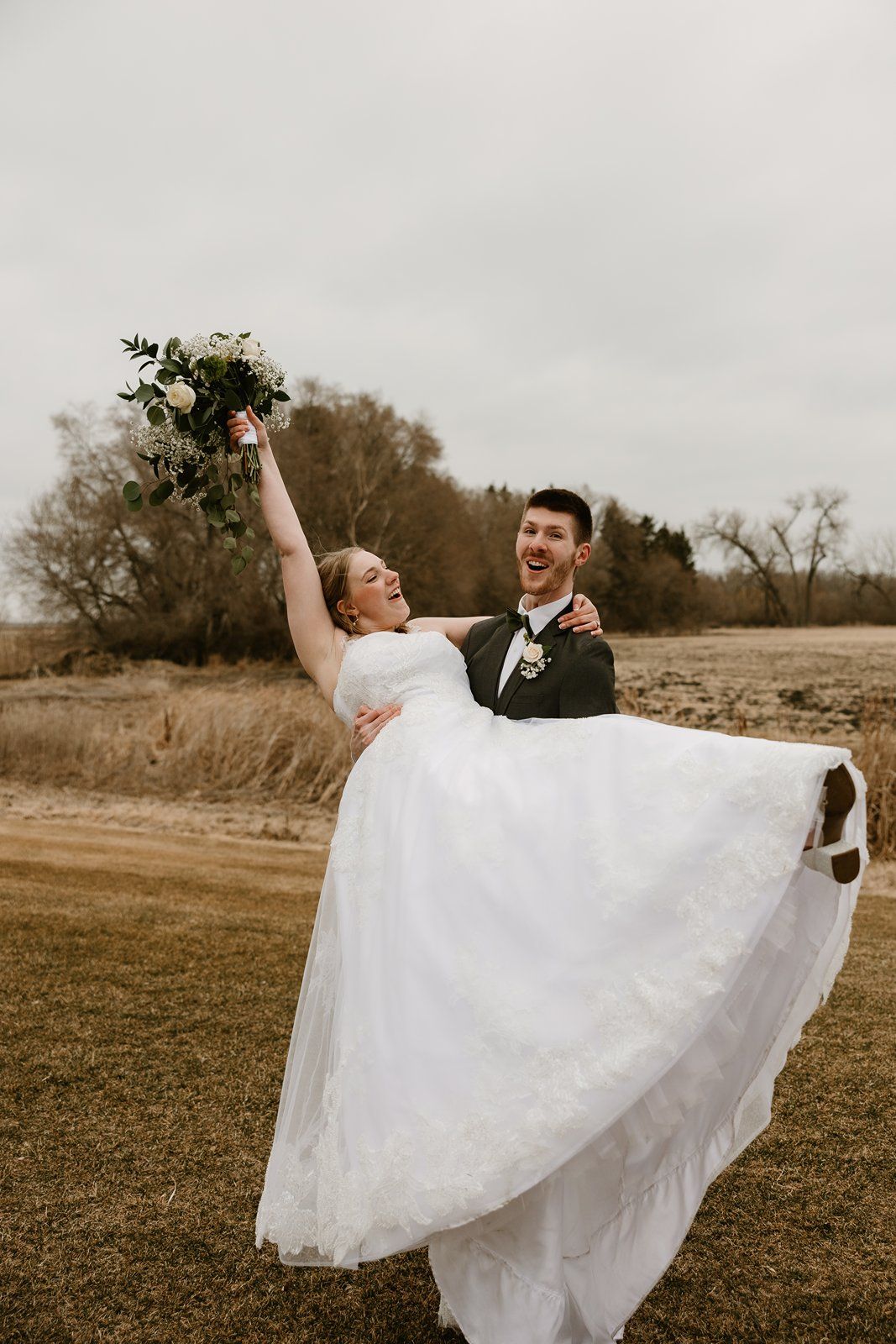 A joyful wedding couple spins and dances together in an open field on an overcast day.