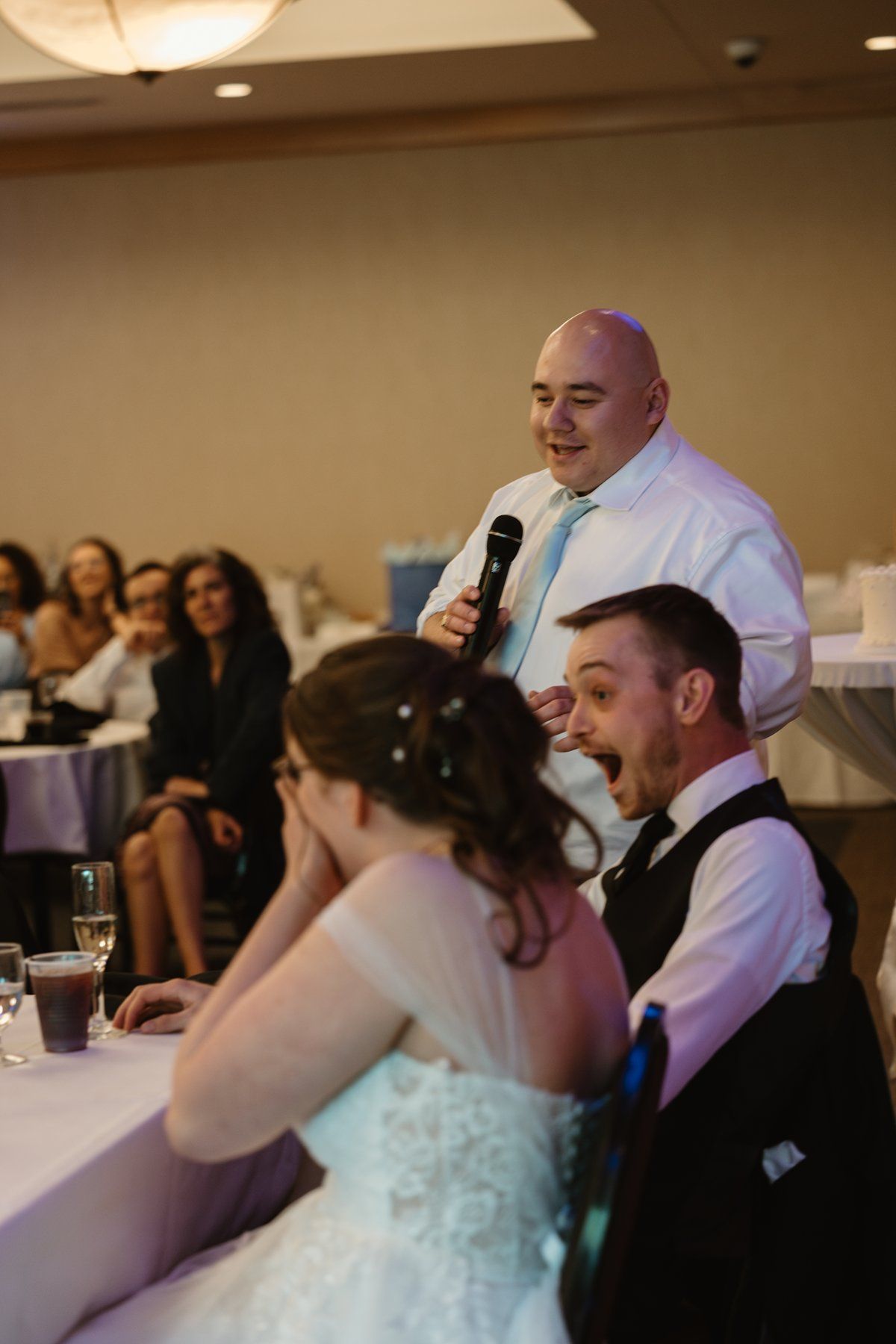 A speaker addresses the newlyweds and guests during a wedding reception dinner in a banquet hall.
