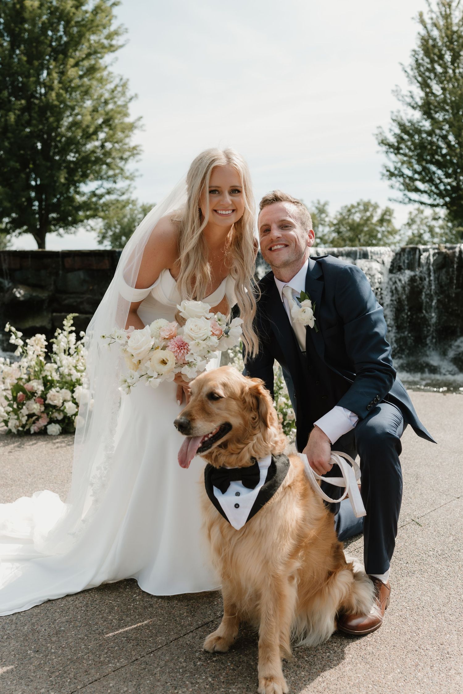 A wedding couple poses with their Golden Retriever wearing a bow tie tuxedo in front of a waterfall backdrop.