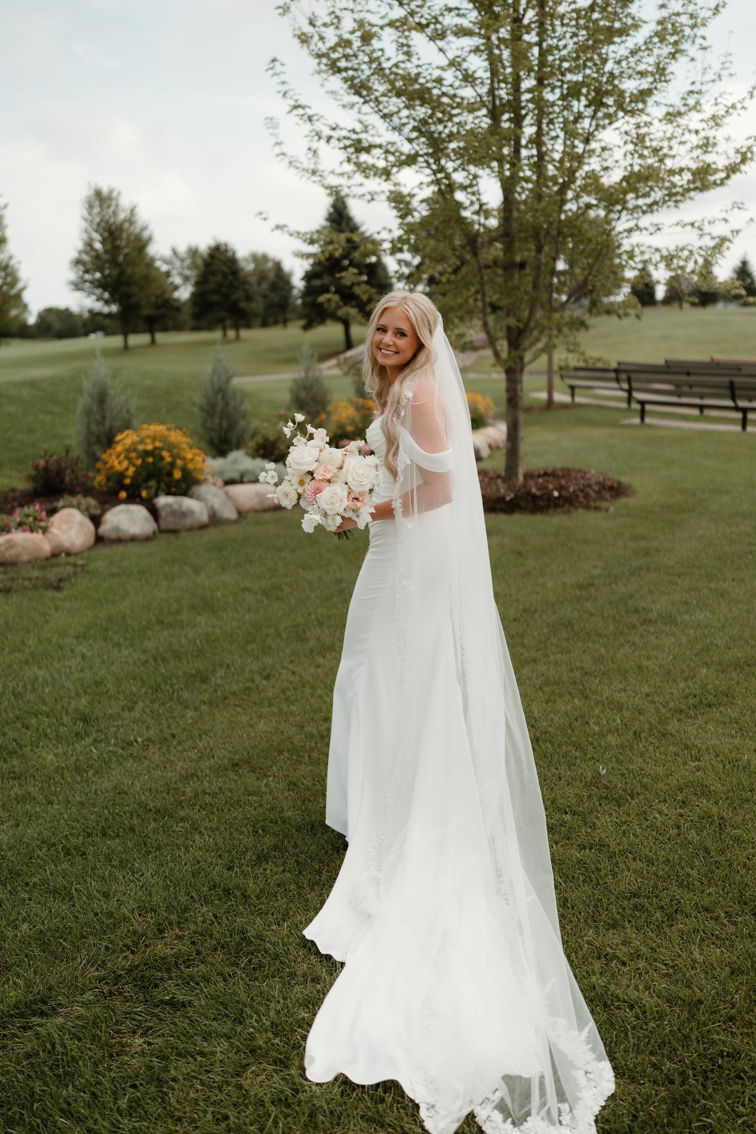 A bride in a flowing white wedding dress stands in a lush green field with trees and landscaping in the background.