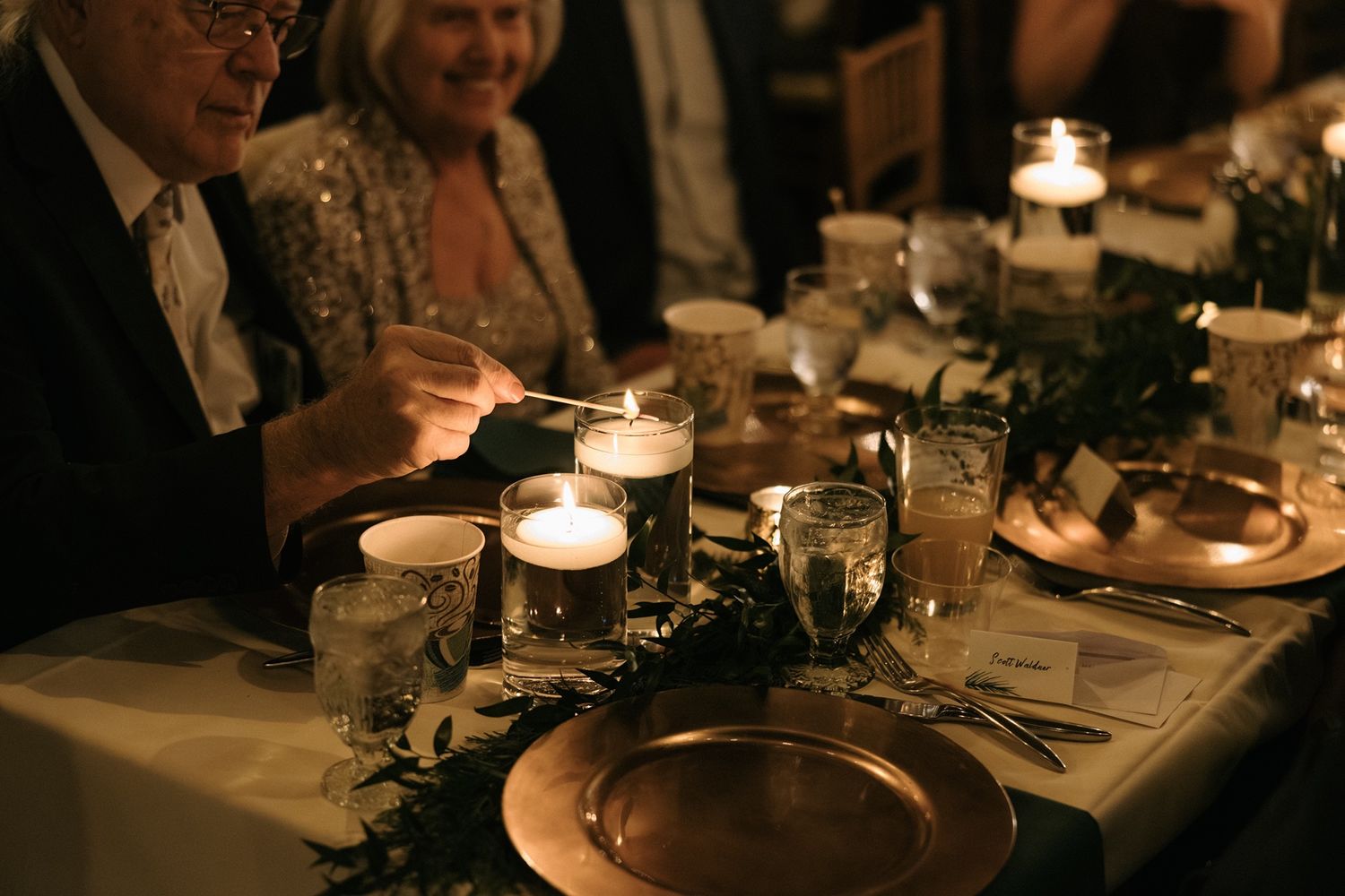 Elegant candlelit dinner table with glowing candles, copper charger plates, and greenery centerpieces at a formal event.