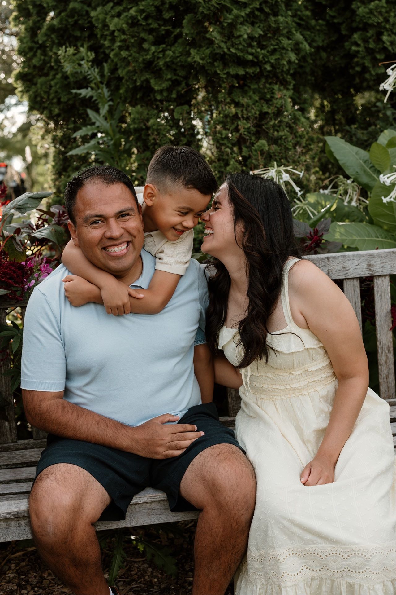 A happy family of three sits on a garden bench, parents smiling as their young son hugs dad from behind.