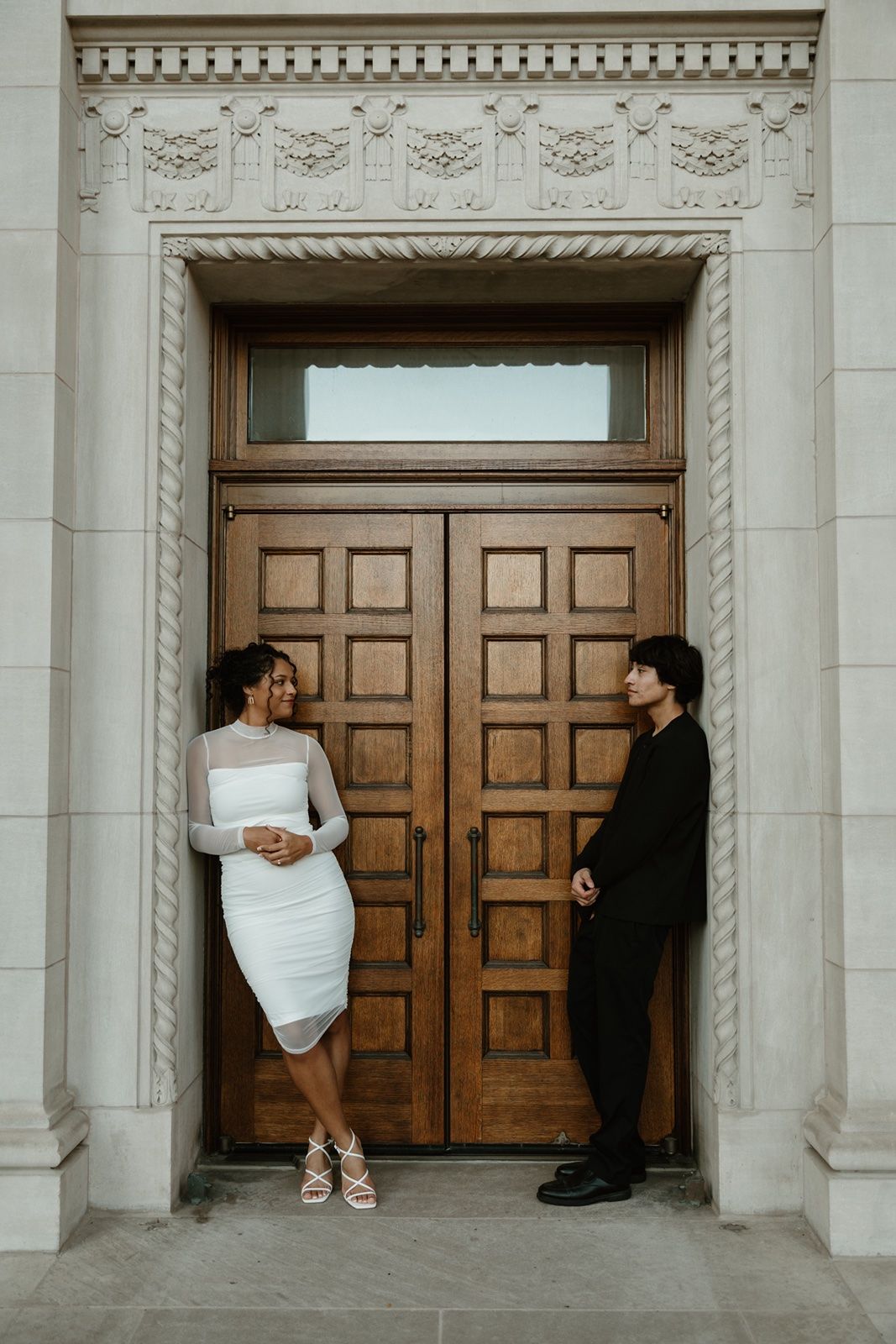 Couple posing in front of elegant wooden double doors at a grand stone building entrance.