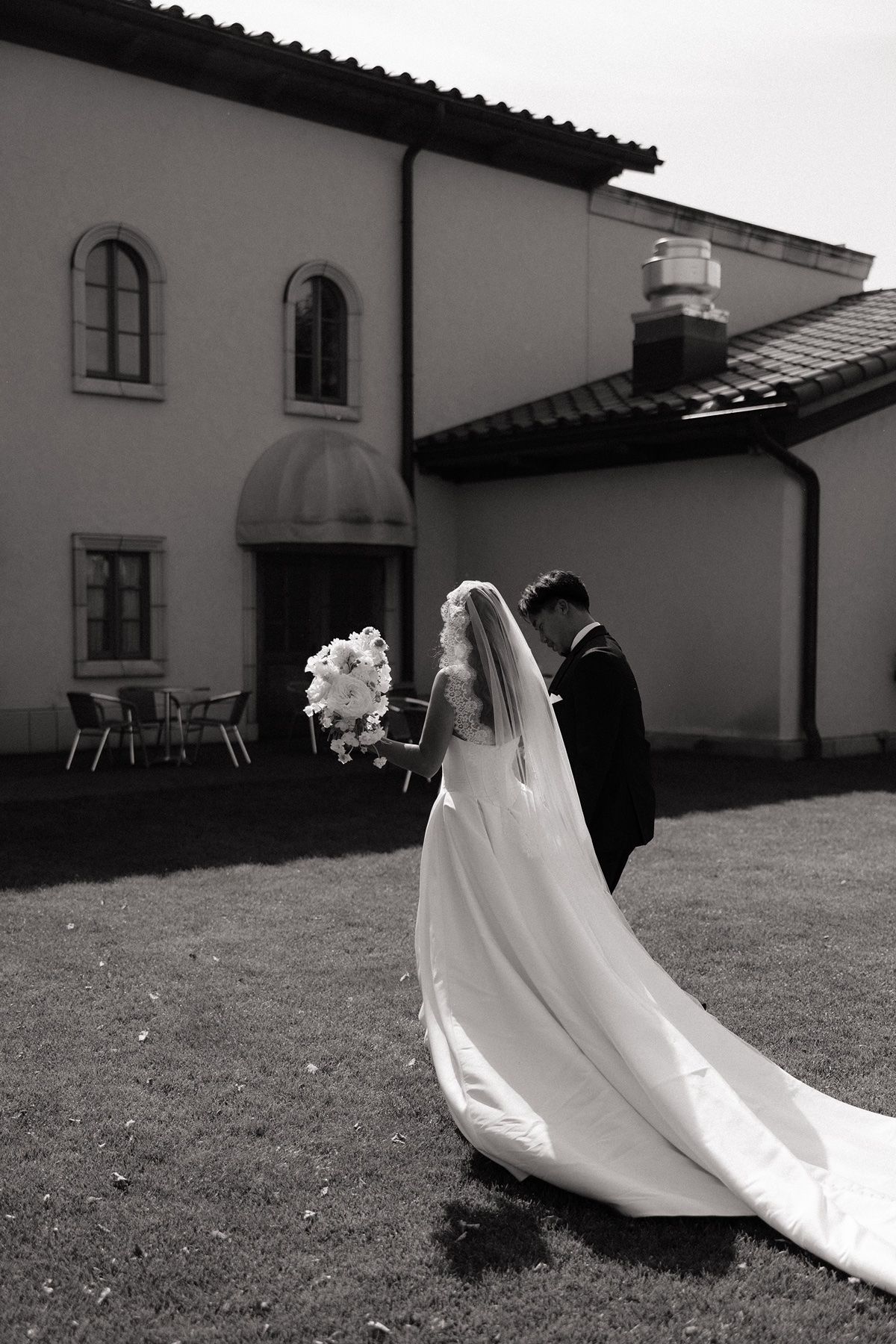 Black and white photo of bride and groom walking toward elegant villa, bride holding bouquet with long veil trailing.