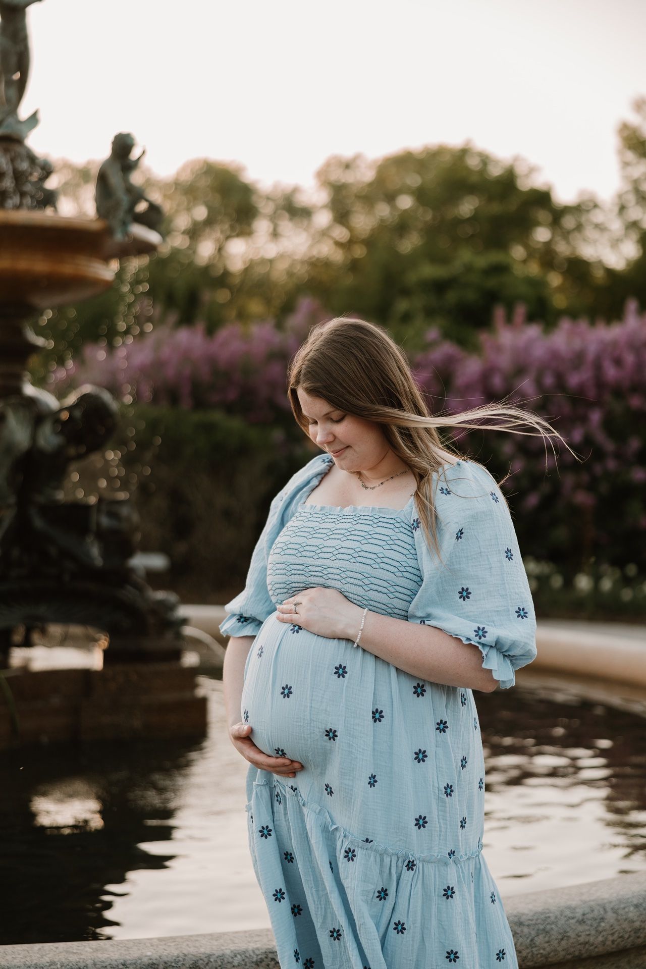 Pregnant woman in blue floral dress cradling baby bump by garden fountain at golden hour.