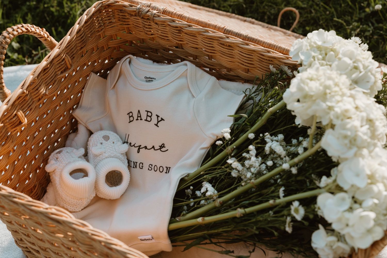 A wicker basket with a white baby onesie, tiny booties, and white flowers, perfect for a baby shower gift.