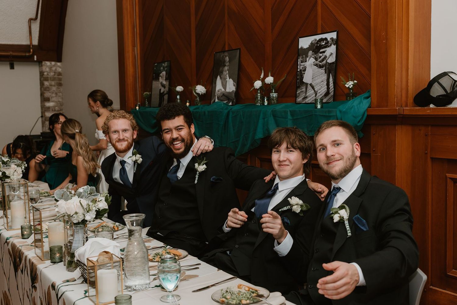 Groomsmen seated at head table smile and raise glasses during wedding reception.