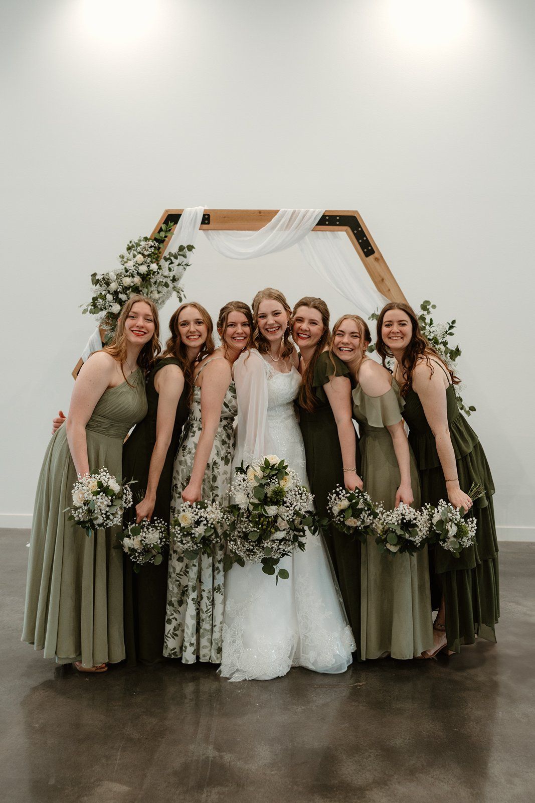 Wedding party wearing sage green bridesmaid dresses pose together with white floral bouquets against neutral wall.
