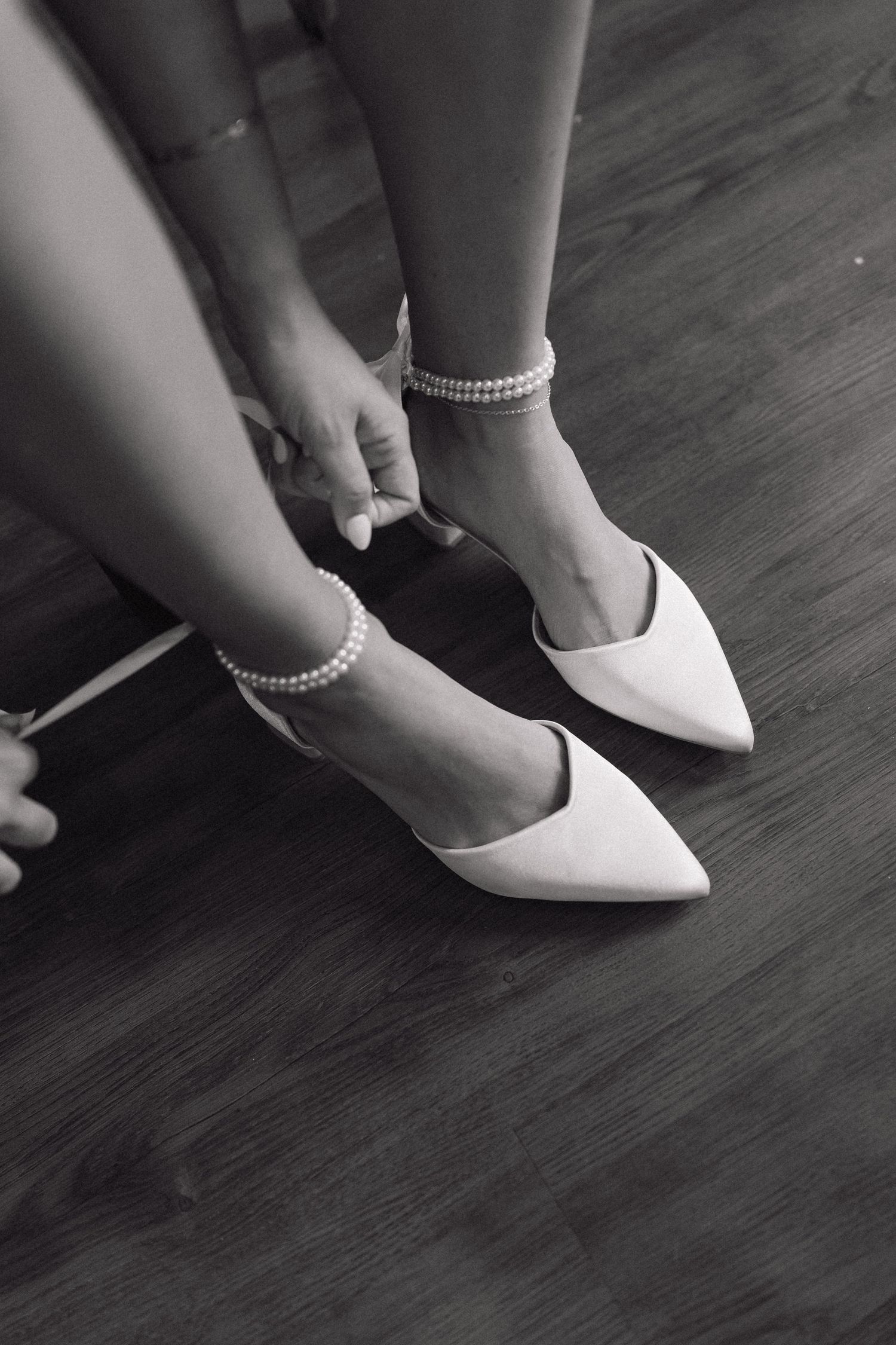 Black and white photo of elegant white pointed flats with pearl ankle straps being put on over bare feet.