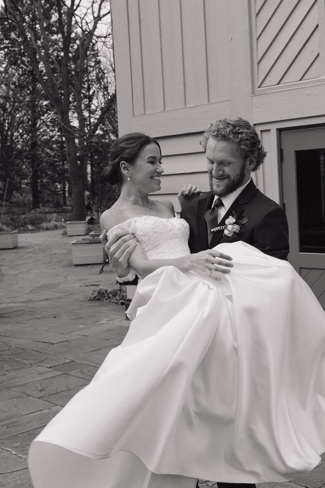 A black and white wedding photo of a couple sharing a playful moment outside.