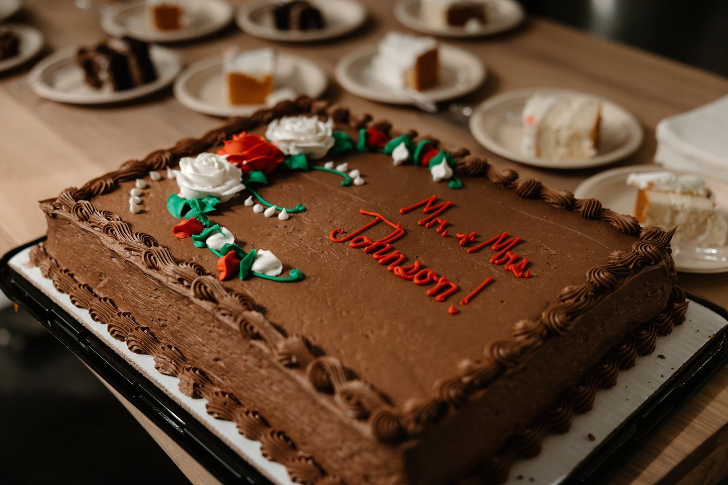 A chocolate sheet cake with red and green frosting decorations sits on a table with other dessert plates in the background.