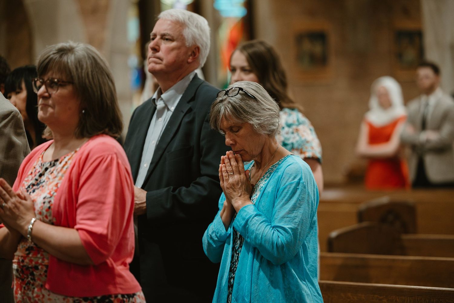 People standing in a church pew during a service, with someone wearing a coral pink blouse and another in turquoise.