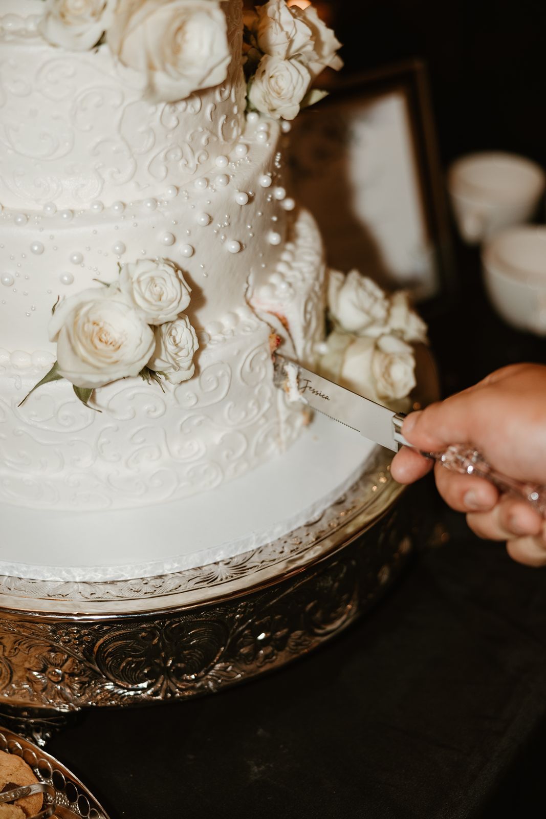 Close-up of white wedding cake with sugar roses being cut on ornate silver stand.