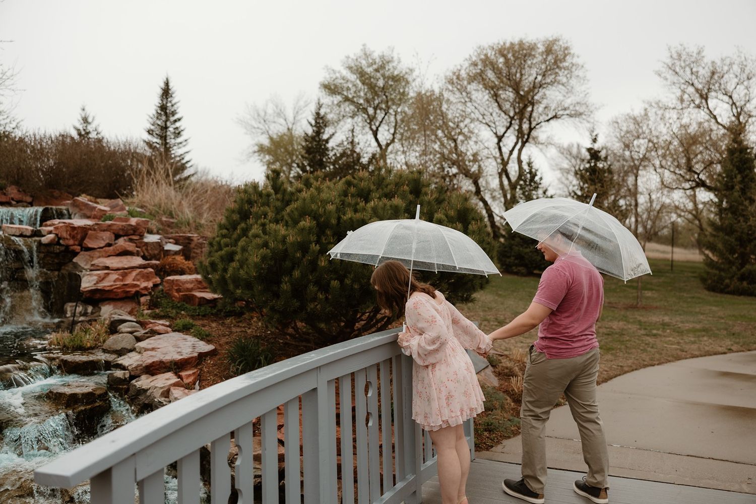 Two people with umbrellas stand on a deck overlooking a landscaped area with stones and trees on an overcast day.