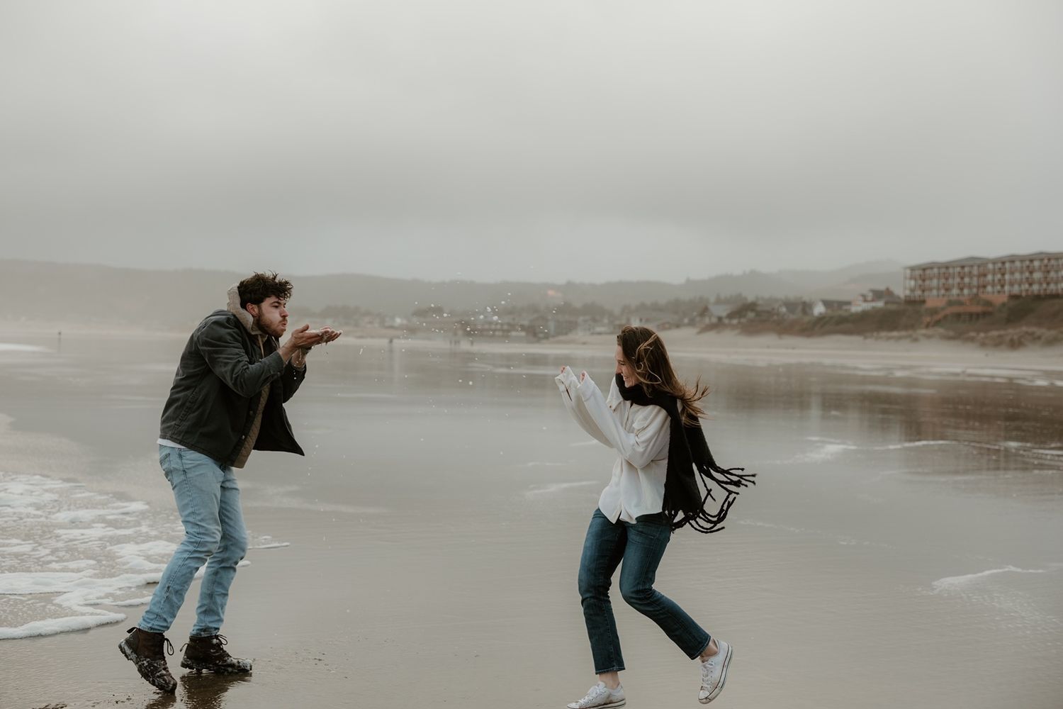 Couple playfully dancing on a misty beach with ocean waves and coastal buildings in the background.