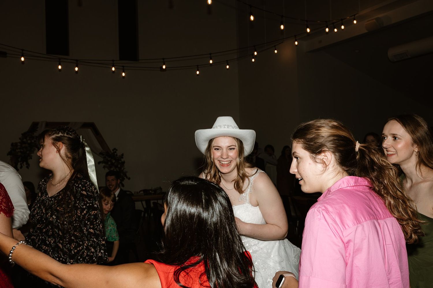 People dancing and socializing at a dimly lit party venue with string lights visible in the background.