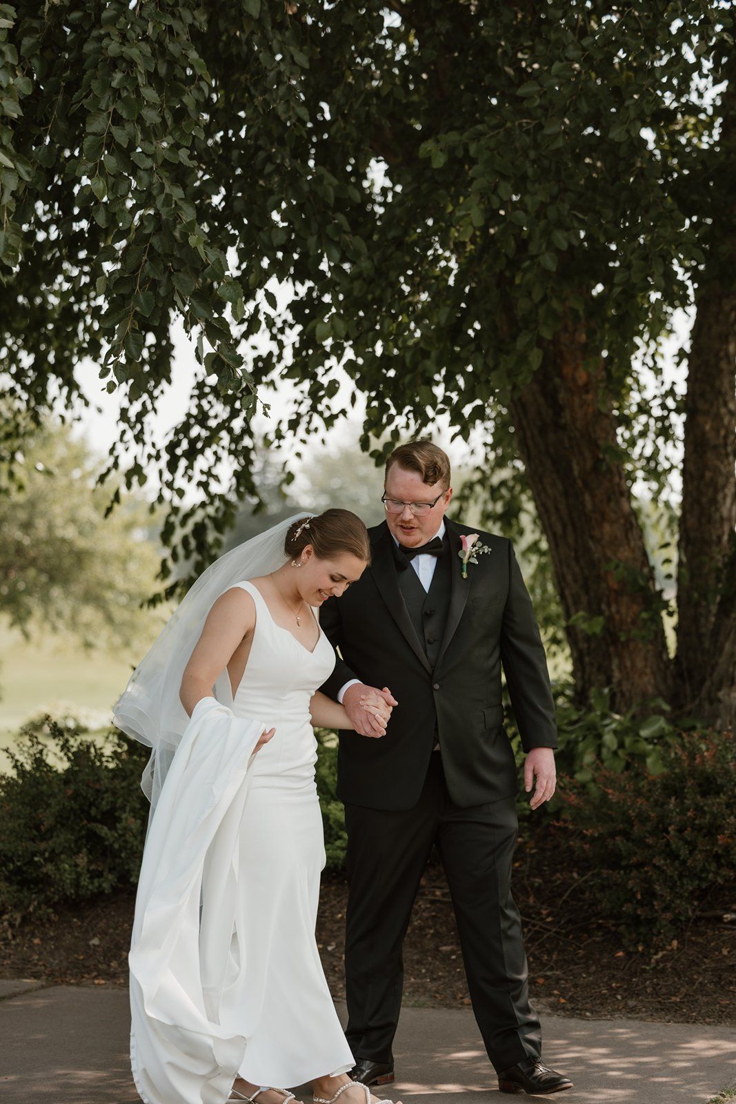 A couple in wedding attire walking together on a path under tree branches on their special day.