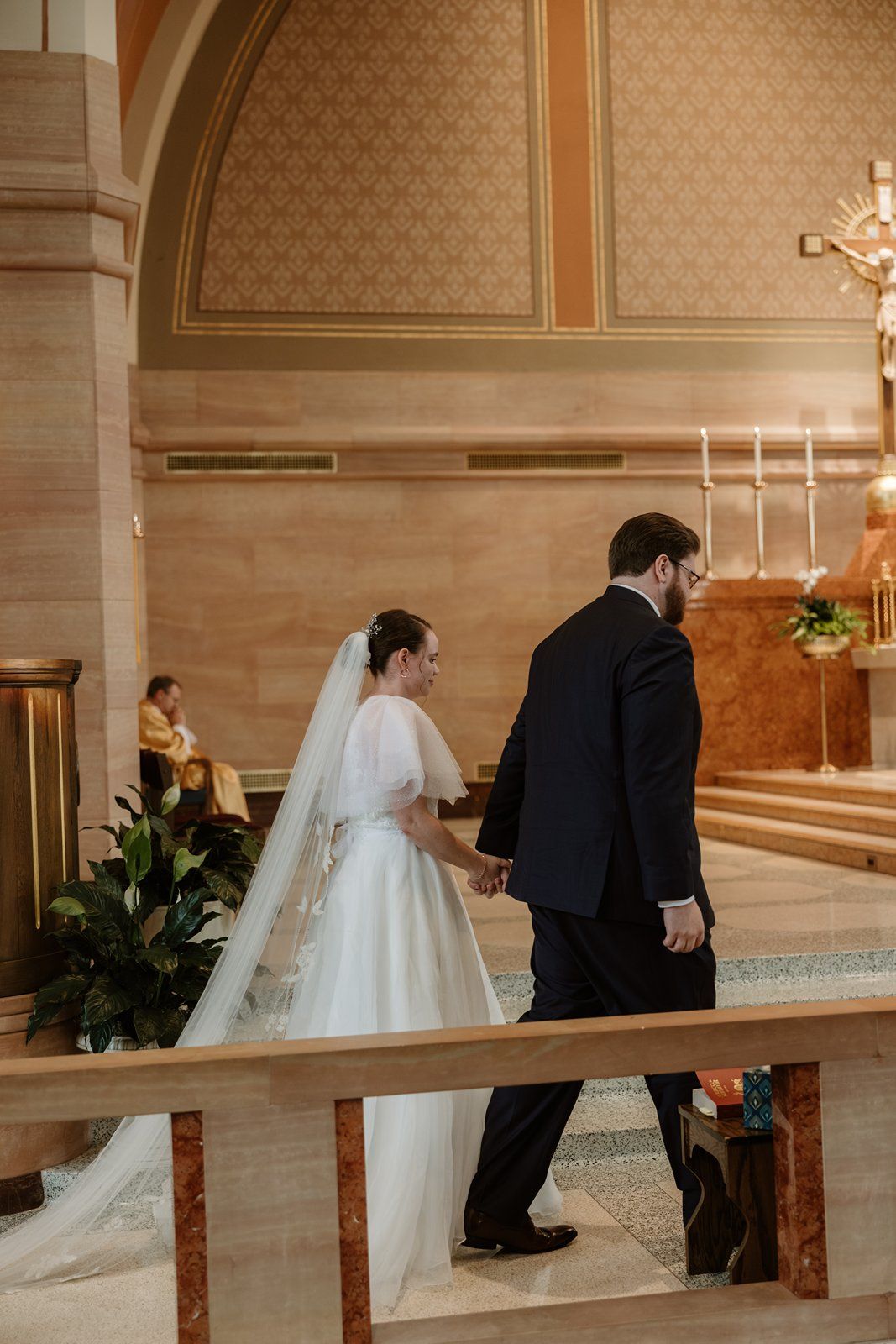 Bride and groom kneel at church altar during wedding ceremony in elegant sanctuary with patterned wallpaper.